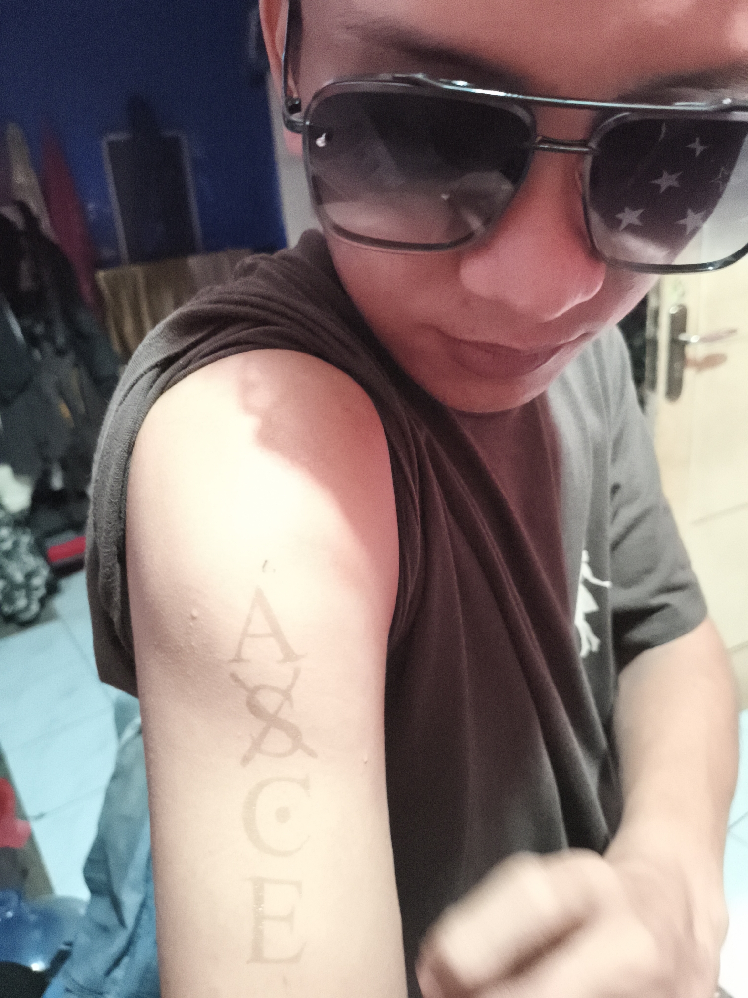 A person wearing sunglasses and a gray t-shirt is facing down and to the right of the frame. Their right arm is visible, displaying a temporary tattoo of the name "ACE" with the letter "S" crossed out. The reflection of stars are visible in the sunglasses. The background shows a cluttered room with various items, including clothing and a piece of furniture. The photograph was taken indoors in Depok, Indonesia.