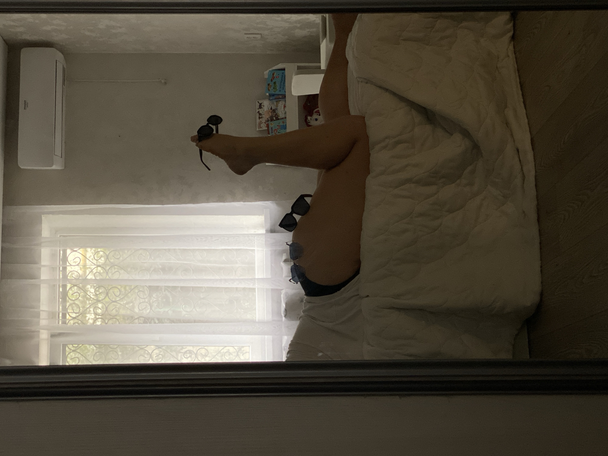 Reflection in a mirror shows a room interior. A person lies on a bed, covered in a white comforter. Their legs extend upwards, one foot holding a pair of sunglasses. Another pair of sunglasses rests on their thigh. A window with sheer patterned curtains is visible. An air conditioning unit is mounted on the wall. Books are visible in the background. The location is potentially Sochi, Russia.