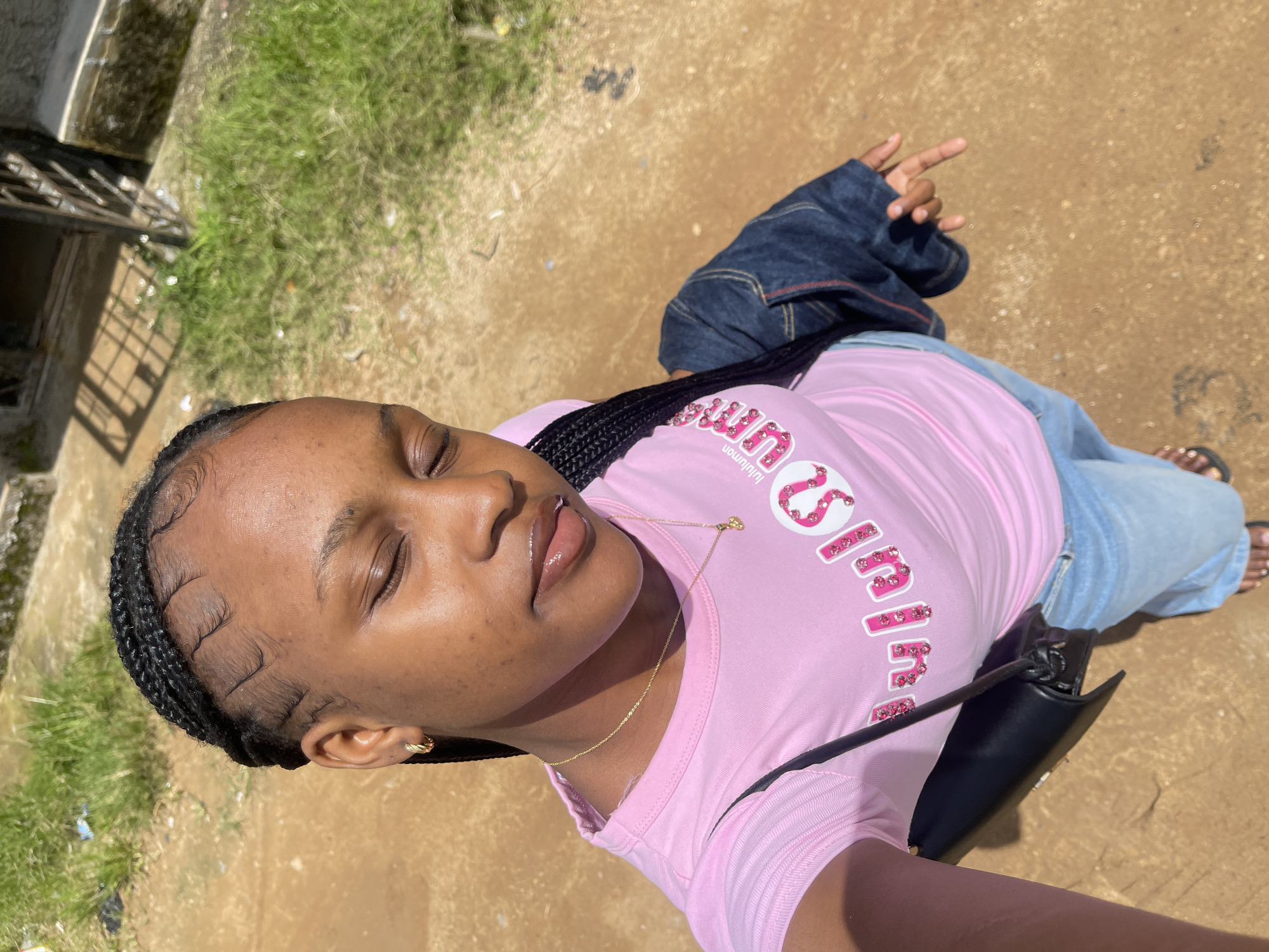 A young Black woman with braided hair and a gold hoop earring poses for a selfie outdoors. She is wearing a pink t-shirt with a glittery design on the front, blue jeans, and a denim jacket draped over her shoulder. A black purse with a strap is slung across her body. Her eyes are closed, and her lips are glossy. The ground beneath her is a mix of dirt and patchy grass, with a building and a metal gate visible in the background. This scene is located in Rafin Par, Nigeria.