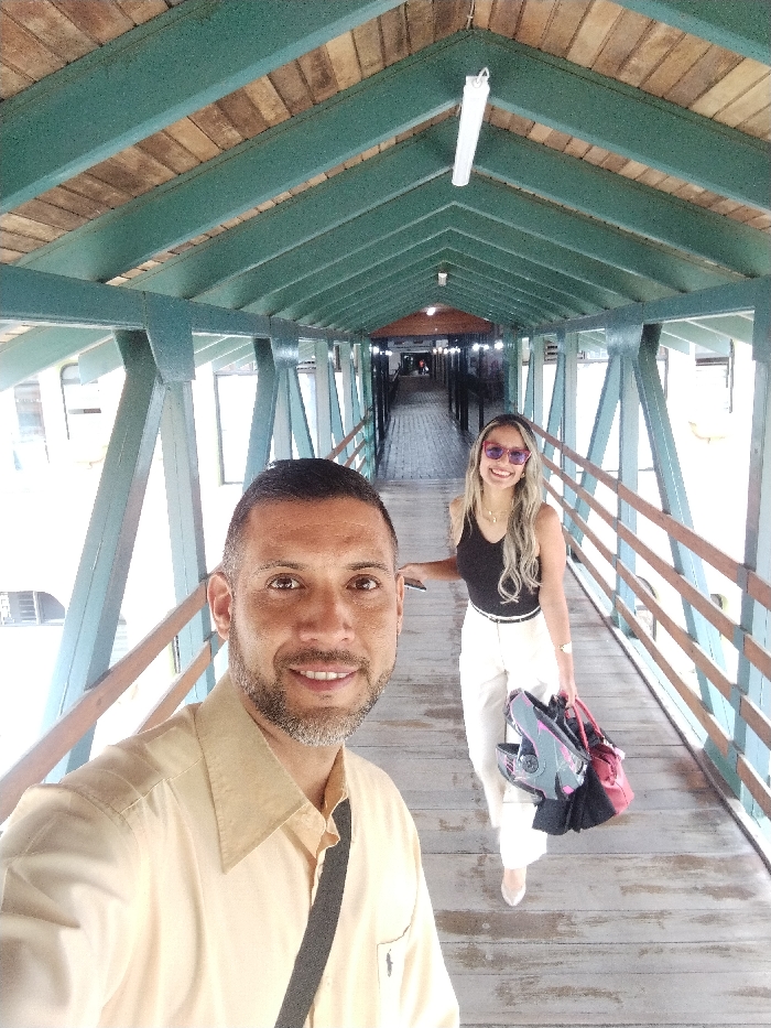A man takes a selfie while walking along a wooden pedestrian bridge with a woman. The bridge has a green-painted frame and wooden railings. The man wears a tan shirt and a black strap across his shoulder. The woman walks ahead, wearing a black top, white pants, and sunglasses, carrying bags. The bridge's ceiling is made of wooden planks. Lights are attached to the ceiling. The background fades into the distance.