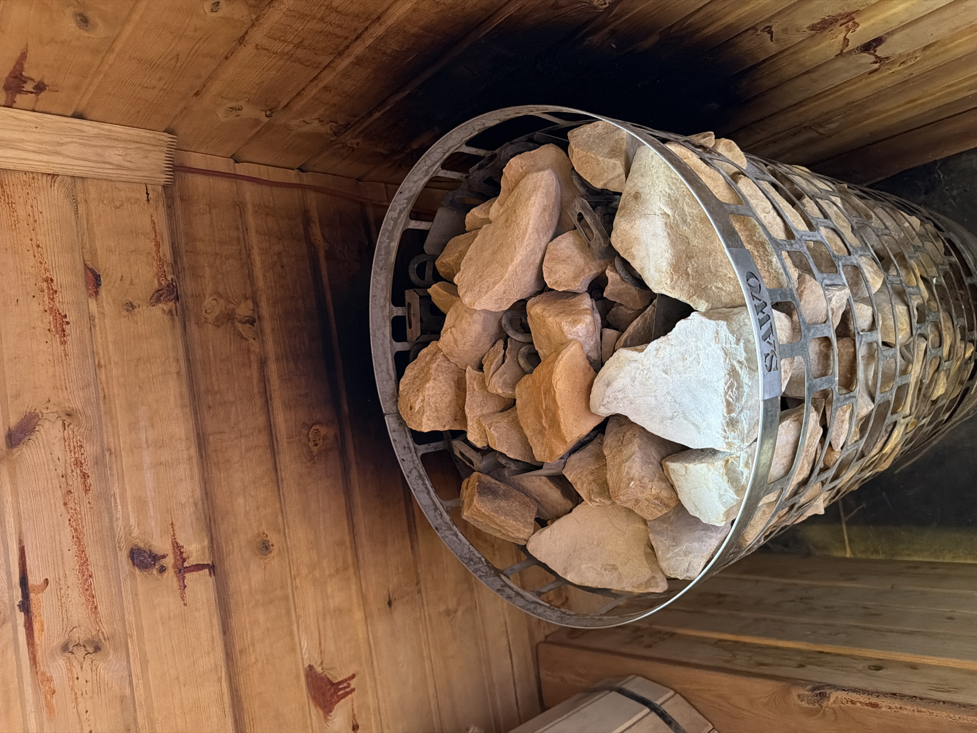 This image shows the interior of a sauna, likely in the city of Semiluki, Russia. The walls and ceiling are constructed of natural wood planks. A central focus of the image is a metal basket filled with heated sauna stones. These stones vary in color from light tan to pale white. The basket itself is cylindrical with a mesh-like exterior and is branded with the word "SAWO". To the left of the basket, a portion of a wooden bucket or container is visible. The overall atmosphere is warm and rustic, typical of a traditional sauna environment.