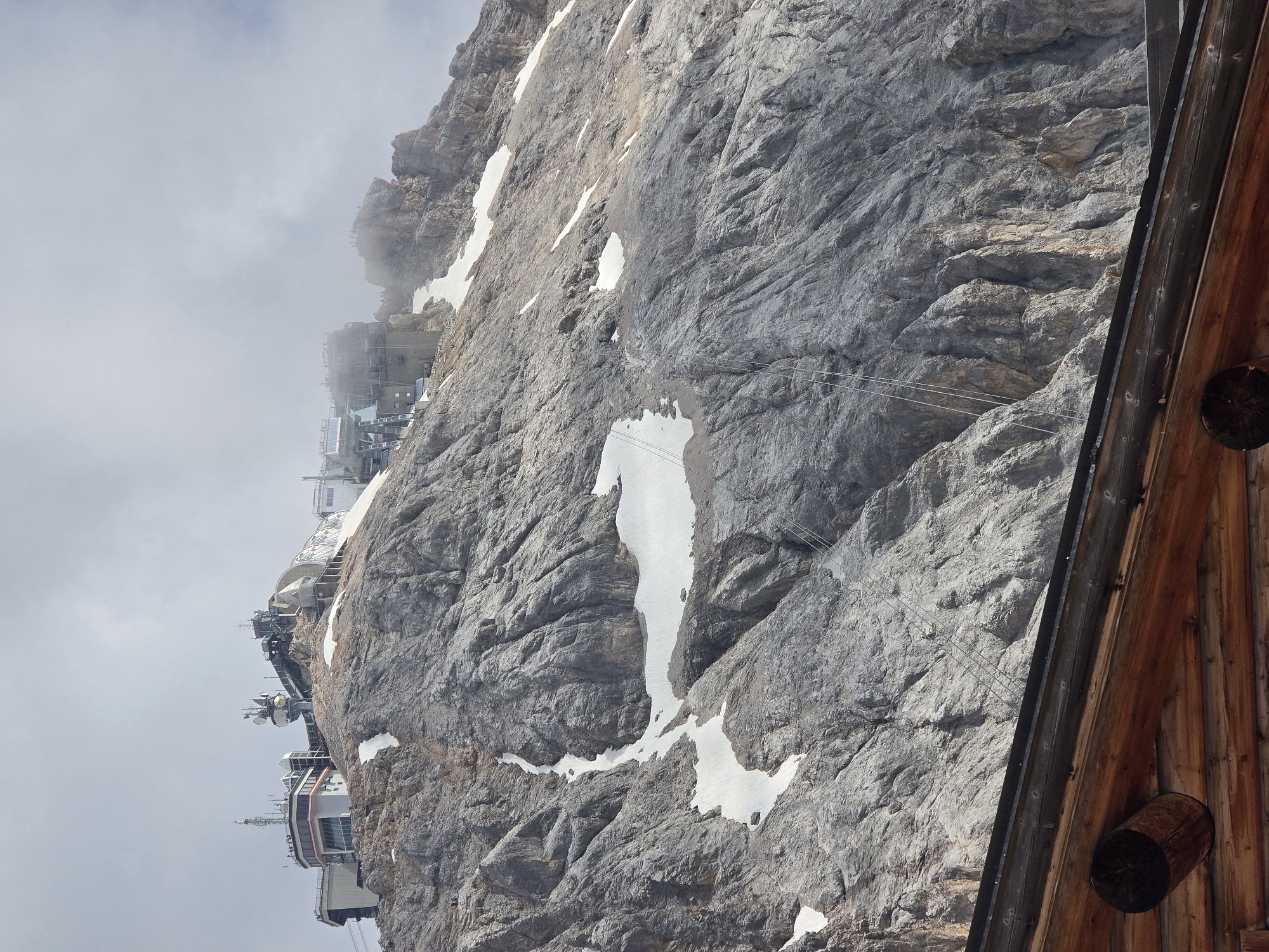The image depicts a steep, rocky mountainside with patches of snow. Several buildings are visible clinging to the cliff face, appearing to be part of a mountain resort or research facility. These structures are multi-leveled and feature modern architectural elements, including large windows and platforms. Cable car lines are also visible ascending the mountain, suggesting a transportation system for visitors.
The weather appears to be overcast, with clouds partially obscuring the upper portions of the mountain and buildings, creating a somewhat misty atmosphere. The overall scene suggests a high-altitude location, likely in a mountainous region. The presence of a wooden structure in the foreground, with visible logs and a circular opening, hints at a rustic or observational element within the otherwise modern mountain infrastructure.
No people are clearly discernible in the image, but the presence of the buildings and cable cars implies that this is a location frequented by humans. The time of day is not definitively indicated, but the ambient light suggests it could be daytime. No visible text is present in the image. The scene evokes a sense of grandeur and the rugged beauty of a mountain environment, juxtaposed with human engineering to access and utilize such a challenging landscape. The location context of Garmisch-Partenkirchen, Germany, aligns with the visual cues of a prominent Alpine mountain.