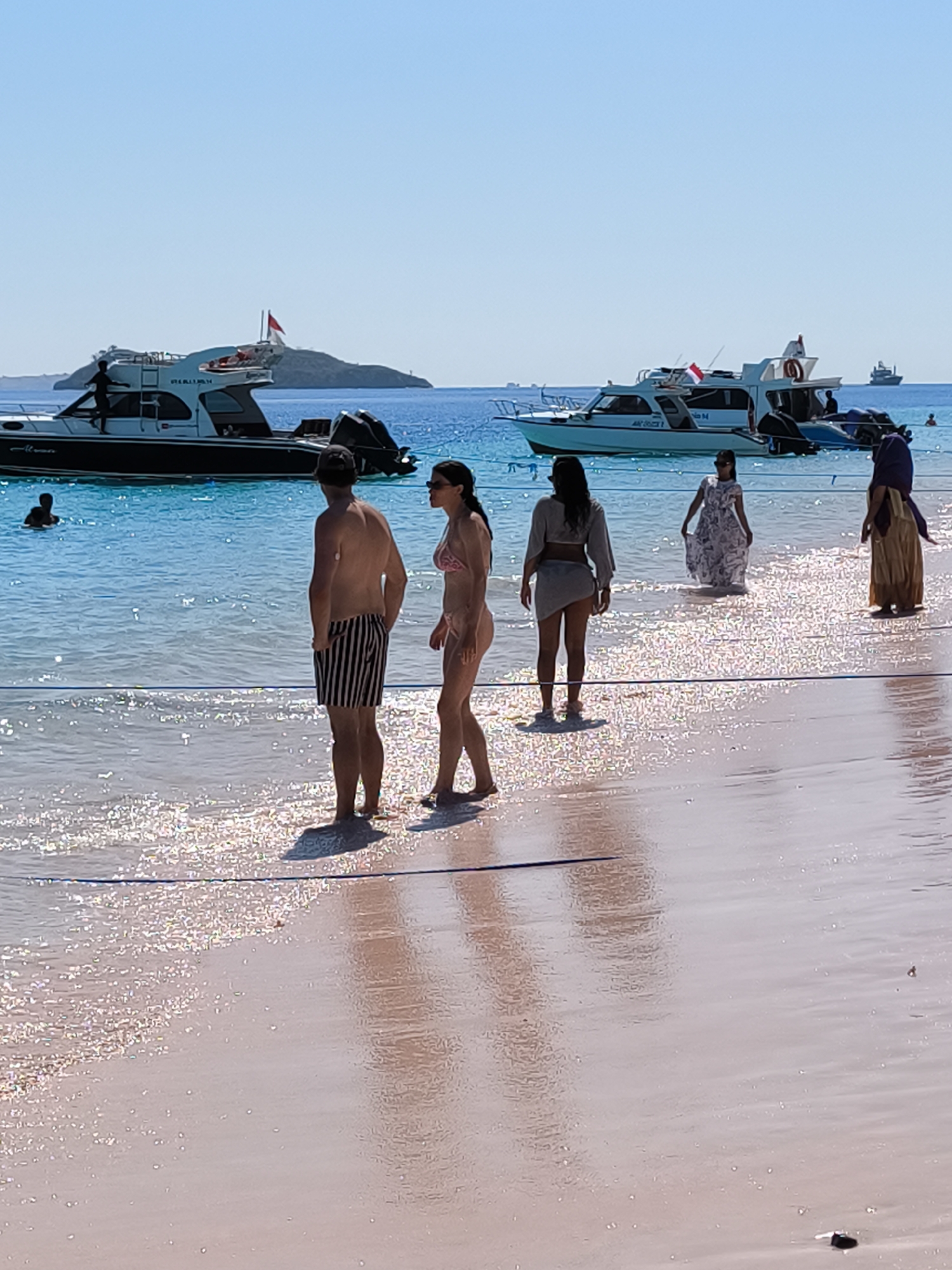 In an outdoor, sunlit scene on a beach in an unknown Indonesian city, several people are visible near the shoreline and in the shallow water. The beach is characterized by pinkish sand with a glistening surface due to the water. The ocean water is clear and turquoise, with small waves lapping at the shore.
In the foreground, a man wearing striped shorts and a woman in a bikini are standing on the wet sand, facing away from the camera. To their right, a woman in a light-colored, patterned dress is walking in the shallow water. Further to the right, another woman, partially obscured, is wearing a long, flowing, tan-colored garment and a head covering.
In the middle ground, several boats are anchored in the water. The closest boat is a white motorboat with a dark hull. In the distance, more boats are visible, as well as a small island or landmass with a distinctive shape. The sky above is clear and bright blue. A person is visible swimming in the water near the boats. The overall impression is a serene beach scene on a sunny day.