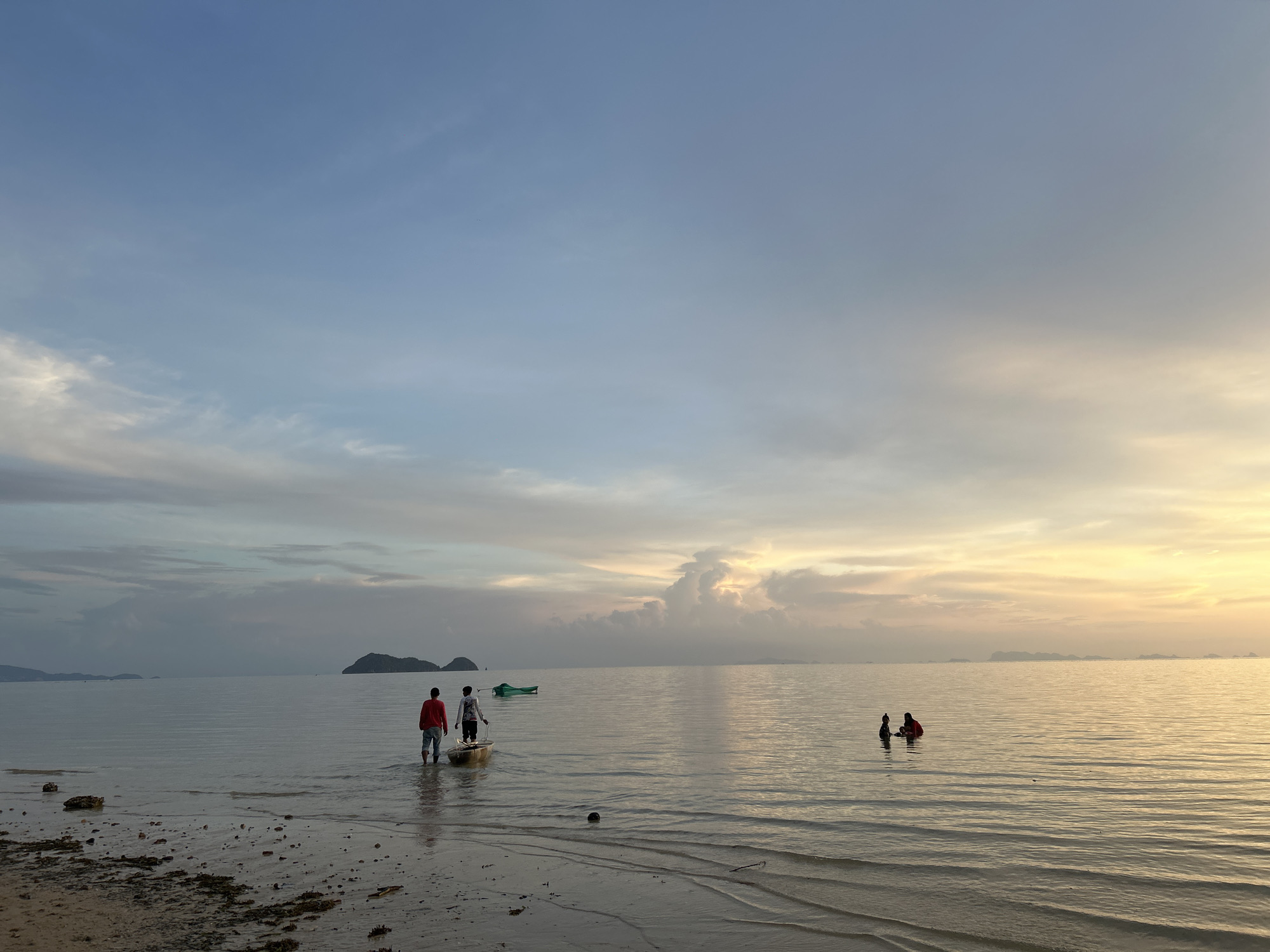 At dusk, two individuals stand knee-deep in the calm ocean, one in a red shirt and the other in a white shirt, facing a small boat. The boat is positioned near the shore, with a green boat floating further out in the water. In the distance, a line of islands emerges from the horizon under a sky transitioning from soft blues to warm oranges and yellows. The sandy shore in the foreground is wet and appears to be low tide, with scattered debris. This scene depicts a serene evening on the coast of Ko Pha-ngan District, Thailand.