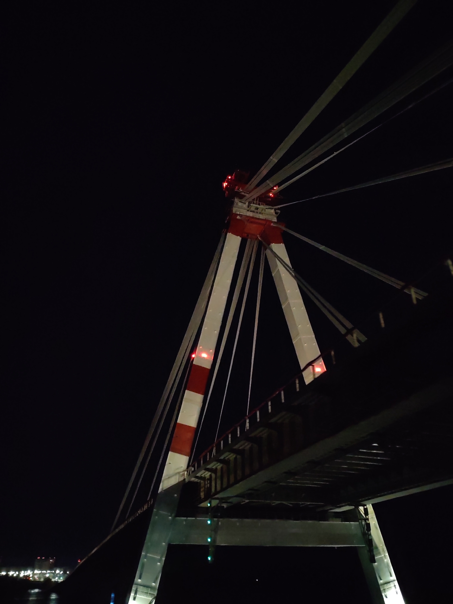 The image depicts a section of the cable-stayed bridge in Cherepovets, Russia, at night. The bridge support tower is prominently displayed, painted with alternating red and white stripes. Cables extend upwards from the tower. The bridge deck is visible, along with supporting steel structures. In the distance, lights from buildings and infrastructure are visible. The scene is dark, with artificial lights illuminating the bridge structure.