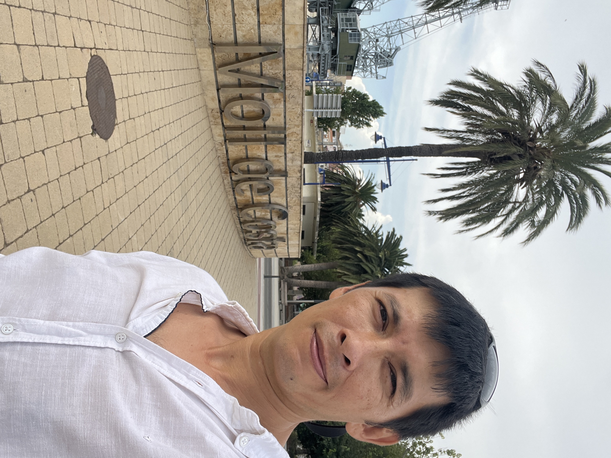 A man wearing a white open-necked shirt takes a selfie with a palm tree and the "Moll de Costa" sign in the background in the city of Tarragona, Spain. The man's face is in the foreground, with sunglasses perched on his head. Behind him, a large sign with metallic letters reads "Moll de Costa," referring to the waterfront area. A tall palm tree is visible to the left of the sign, and further in the background, industrial cranes and buildings can be seen. The ground is paved with interlocking light brown bricks, and a circular manhole cover is visible in the foreground. The sky is overcast.
