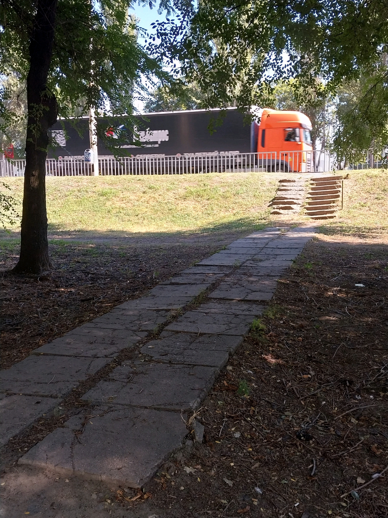 The image depicts a paved pathway in a park-like setting, with a truck visible in the background. The pathway, made of square concrete slabs, appears weathered and is bordered by dry leaves and twigs. To the right of the pathway, a set of concrete stairs ascends a grassy incline. In the background, a large black truck with an orange cab is partially visible, moving along a road behind a white metal fence. The scene is bathed in sunlight, casting shadows on the ground. Trees with green foliage frame the upper part of the image. This location is likely in the city of Dnipro, Ukraine, given the context.