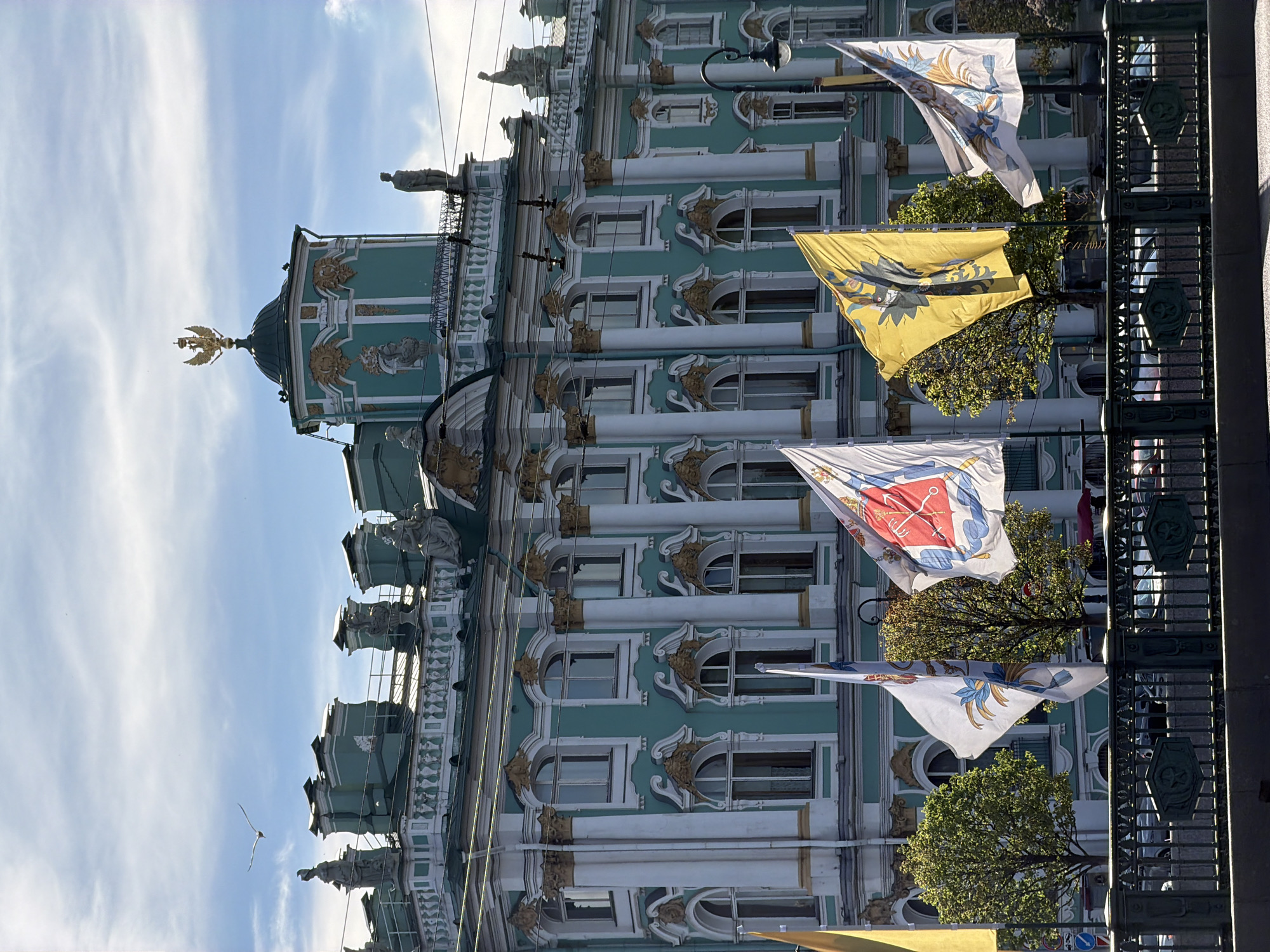 The image captures a view of the Winter Palace in Saint Petersburg, Russia, a prominent landmark on Palace Square. The palace, painted in a distinctive aquamarine color, is adorned with white and gold architectural details, including columns, arches, and decorative sculptures along its roofline. Atop one of the central turrets, a golden double-headed eagle crest gleams under the daylight. In the foreground, several flags are unfurled, adding a splash of color to the scene. Among them, a yellow flag with a black emblem and a white flag with a red crest are prominently displayed. The flags are mounted on poles and appear to be near a body of water, possibly the Moika River or the Neva River, suggested by the presence of a decorative black metal fence in the very front, which might be a bridge railing. Trees with vibrant green foliage are scattered behind the flags, some appearing to be meticulously pruned. The sky above is a bright blue with wispy white clouds, and a single bird is visible in flight. The overall impression is one of grandeur and historical significance, typical of Saint Petersburg's imperial architecture.