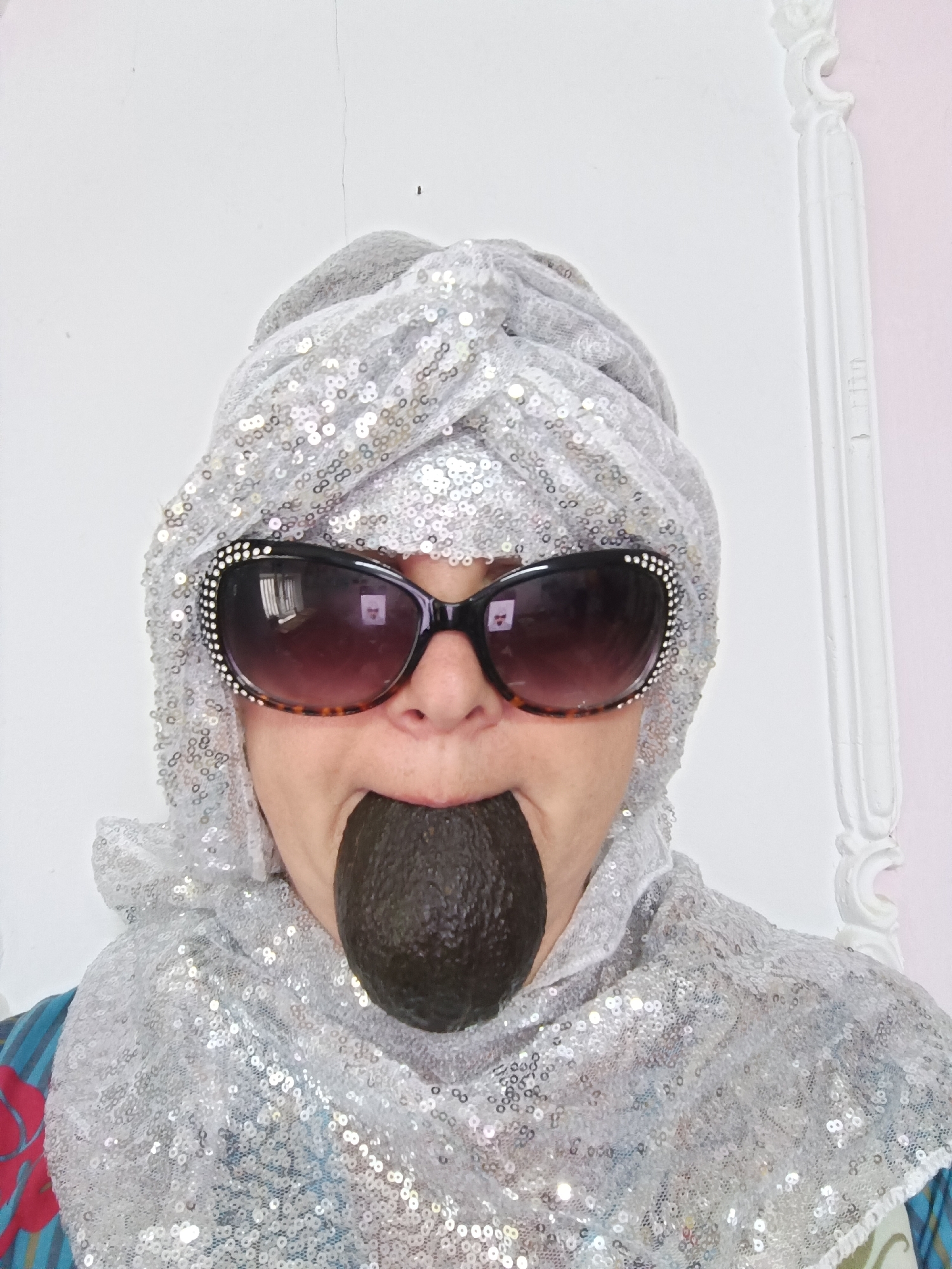 A person, possibly female, is pictured head-on, wearing a silver sequined turban and matching shawl. Oversized, dark-lensed sunglasses with a tortoiseshell pattern and rhinestone embellishments adorn their face. Their mouth is open, with a dark, oval-shaped object, possibly an avocado, held within. The background is a plain white wall with some subtle texture, and a white decorative molding is visible on the right side. The image appears to be a selfie taken indoors.
