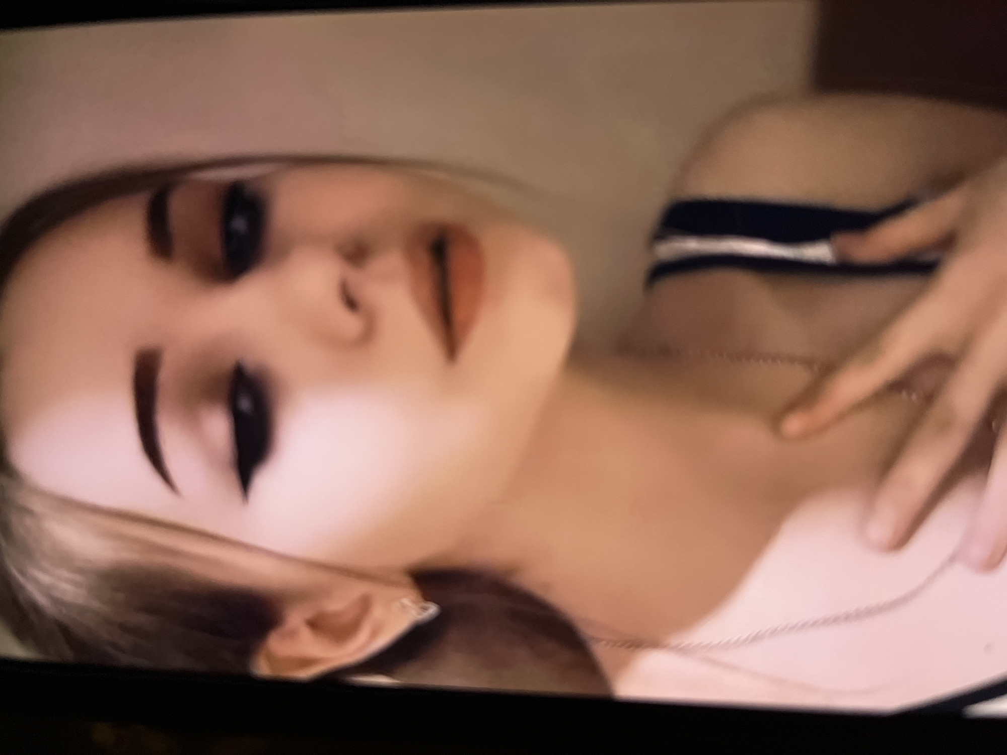 A portrait-style image, likely a photograph of a screen, features a young woman from the shoulders up. Her head is tilted, and she looks directly towards the viewer. She has light-colored hair, prominent dark eyeliner, and well-defined eyebrows. Her lips are painted with a warm, possibly reddish-orange, shade. A small nose piercing is visible on her left nostril. She wears a light-colored top, with a black and white striped strap visible over her right shoulder, suggesting a bra or tank top strap. A thin chain necklace rests on her chest, and a ring adorns a finger on her right hand, which is positioned near her left shoulder. The background is softly blurred and appears to be a uniform light tone, indicating an indoor setting. Her expression is direct and composed. No text is visible in the image.