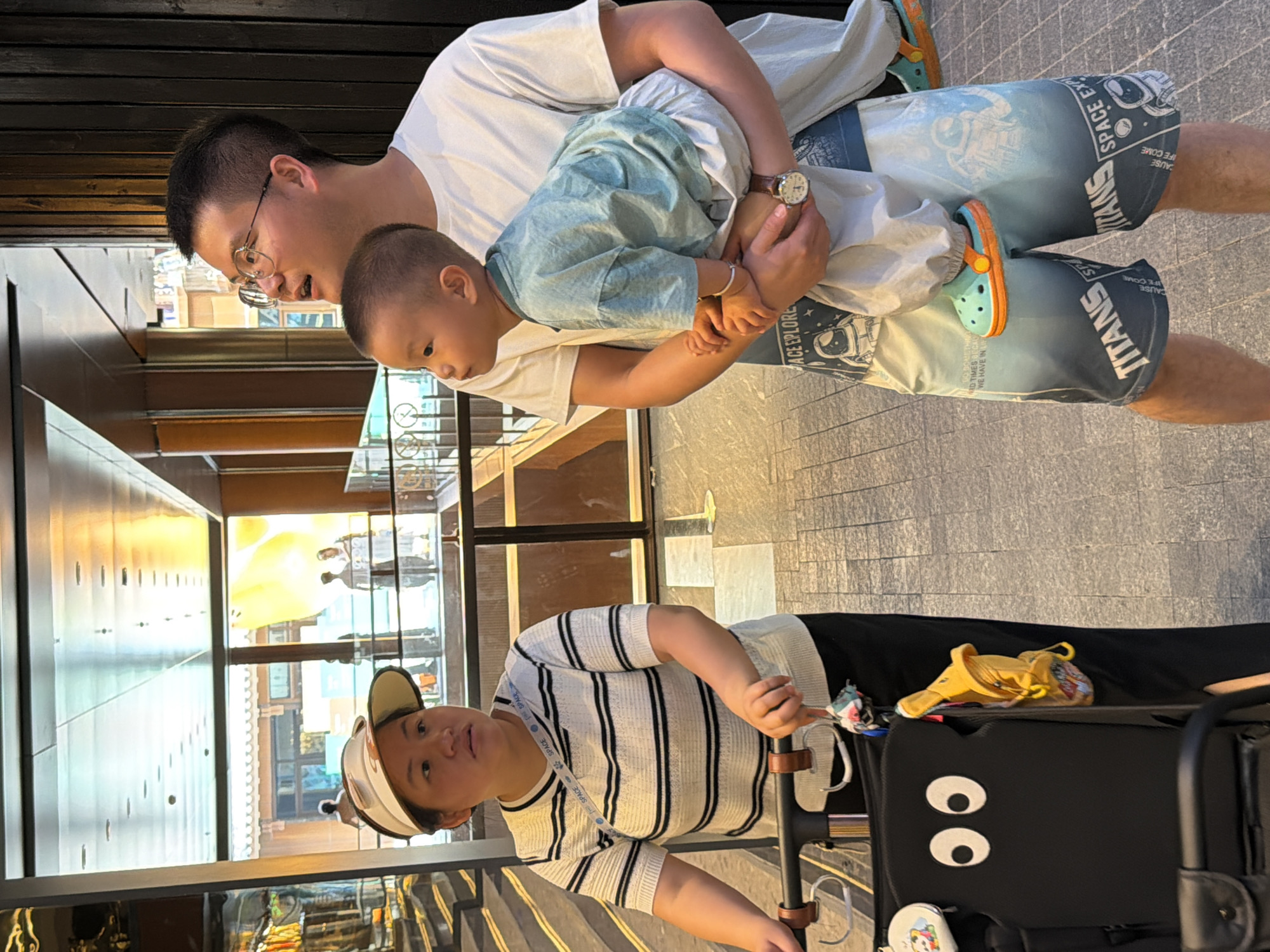 In the city of Yuzhong District, China, a man wearing glasses and a white t-shirt is holding a young child. The child is wearing a light blue t-shirt and light-colored pants. The man is also wearing shorts with a space-themed print. A woman wearing a visor and a striped t-shirt is standing next to a stroller, looking to the left. The stroller is black and has two large white eyes depicted on the backrest. A yellow toy is attached to the stroller's handle. The background shows a modern interior with large windows, illuminated ceiling lights, and wooden paneling. The floor is paved with patterned tiles.