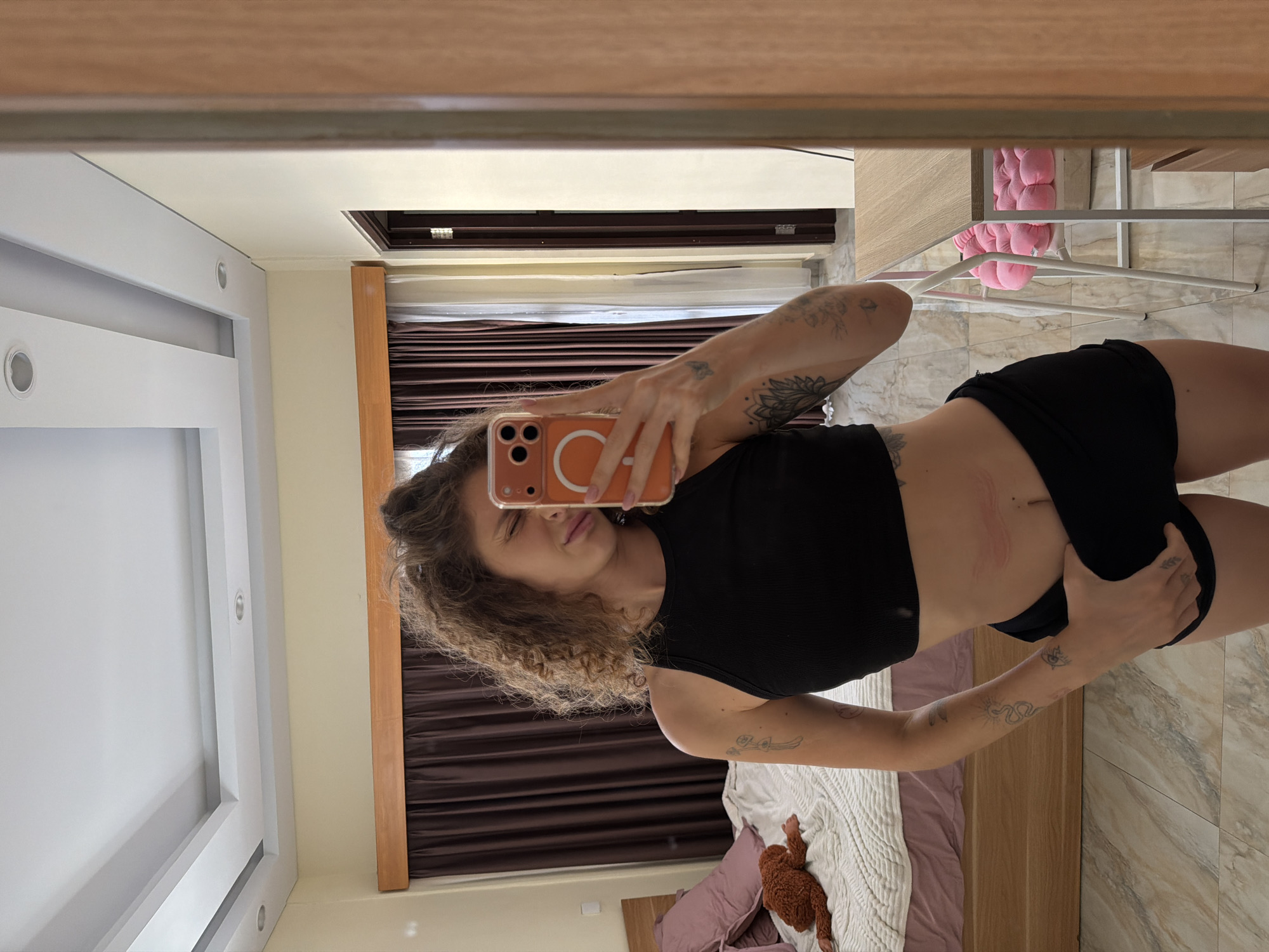A young woman with curly brown hair is captured in a mirror selfie, rotated 90 degrees clockwise, in what appears to be a bedroom or hotel room in North Kuta, Indonesia. She is wearing a black sleeveless crop top and black shorts. Her face is partially obscured by her orange-cased smartphone, which features three camera lenses and a visible circular magnetic attachment. She pouts playfully for the camera, while her other hand rests on her shorts.
Visible tattoos adorn her arms, including a sun and snake design on her right forearm and floral patterns on her left upper arm. A reddish mark is present on her lower abdomen.
The background, reflected in the mirror, shows a bed with a white textured blanket, purple pillows, and a wooden headboard. A brown plush toy is situated on the bed. The room features light-colored walls, a white ceiling with recessed lighting, and dark brown slatted blinds or curtains over a window. The floor is covered in light-colored, patterned tiles, possibly marble. To the right of the reflection, a white metal frame structure with a pink fluffy cushion and light wooden accents are visible. The scene is well-lit, suggesting an indoor setting during the day or a brightly lit interior. No discernible text is visible in the image.
