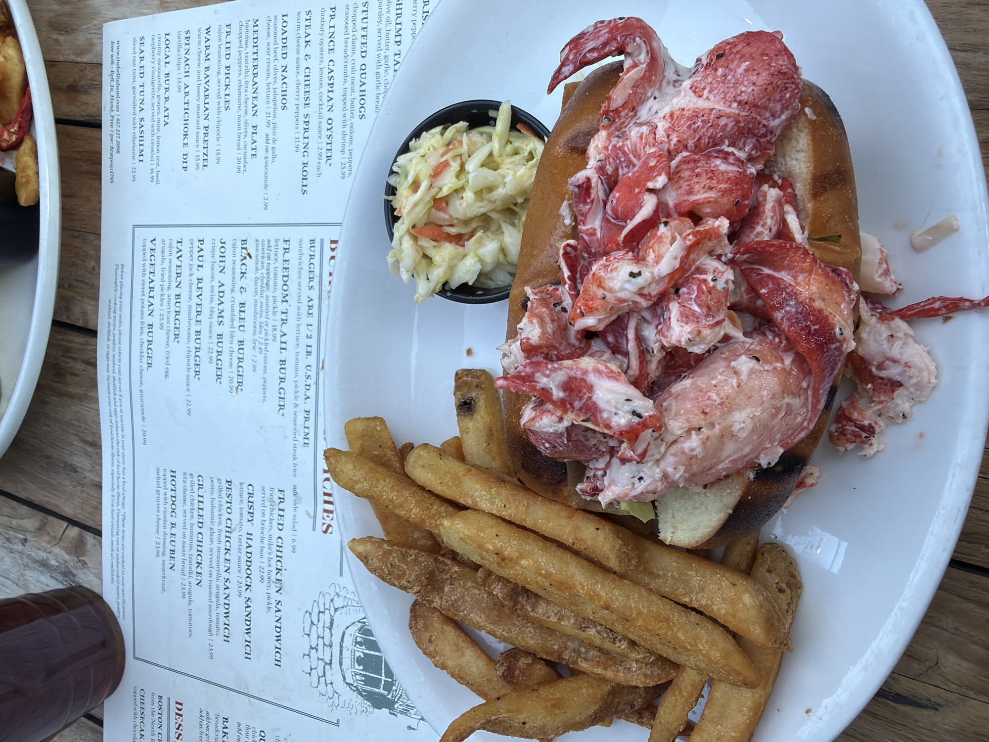 A close-up, high-angle shot shows a lobster roll and french fries on a white plate, set against a blurry background of a menu and a glass of dark liquid. The lobster roll is split in half, revealing a generous filling of lobster meat mixed with mayonnaise and seasoning, served on a toasted bun. Next to it, a mound of golden-brown, thick-cut french fries is piled high. A small bowl of coleslaw sits between the fries and the lobster roll. The menu, visible in the upper portion of the image, lists various seafood dishes and sandwiches. The setting appears to be a restaurant in Boston, Massachusetts, as indicated by the menu item "BOSTON COLD LOBSTER ROLL". The lighting is natural, creating soft shadows and highlighting the textures of the food. No people are visible in the frame.