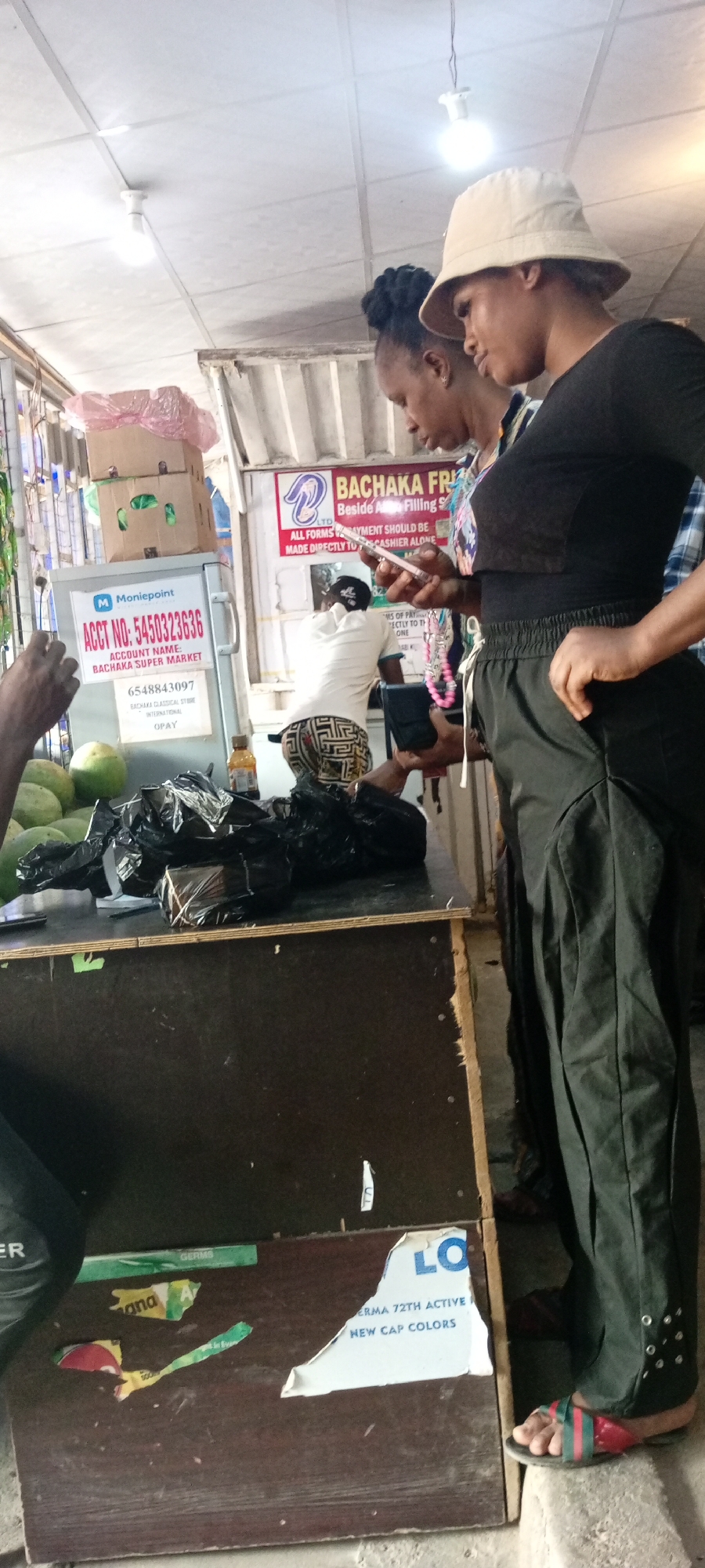The image shows three people at what appears to be a market stall in Aviation Quarters, Nigeria. In the foreground, a person's arm is visible on the left, reaching towards a pile of green fruits, possibly melons, and some items wrapped in black plastic bags on a wooden counter.
Two women are standing behind the counter, both looking down. One woman, wearing a black top and dark green pants with decorative eyelets at the ankle, is holding a smartphone and appears to be engaged in a transaction or interaction on it. The other woman, in a striped top and a light-colored bucket hat, stands next to her. Behind them, another person in a white shirt and patterned pants is partially visible, seemingly working at the stall.
Various signs are visible on and around the stall. One sign displays "Moniepoint" and an account number: "ACCT NO: 5450323636" for "BACHAKA SUPER MARKET". Another sign reads "BACHAKA FRIL LTD Beside A Filling S" and "ALL FORMS PAYMENT SHOULD BE MADE DIRECTLY TO CASHIER ALONE". Other visible text includes "OPAY", "GERMS", "ana", and "ERMA 72TH ACTIVE NEW CAP COLORS".
The lighting suggests it could be daytime, possibly indoors or under a covered area given the presence of ceiling lights and what looks like a corrugated metal ceiling. The overall scene depicts a bustling marketplace environment where transactions are taking place.