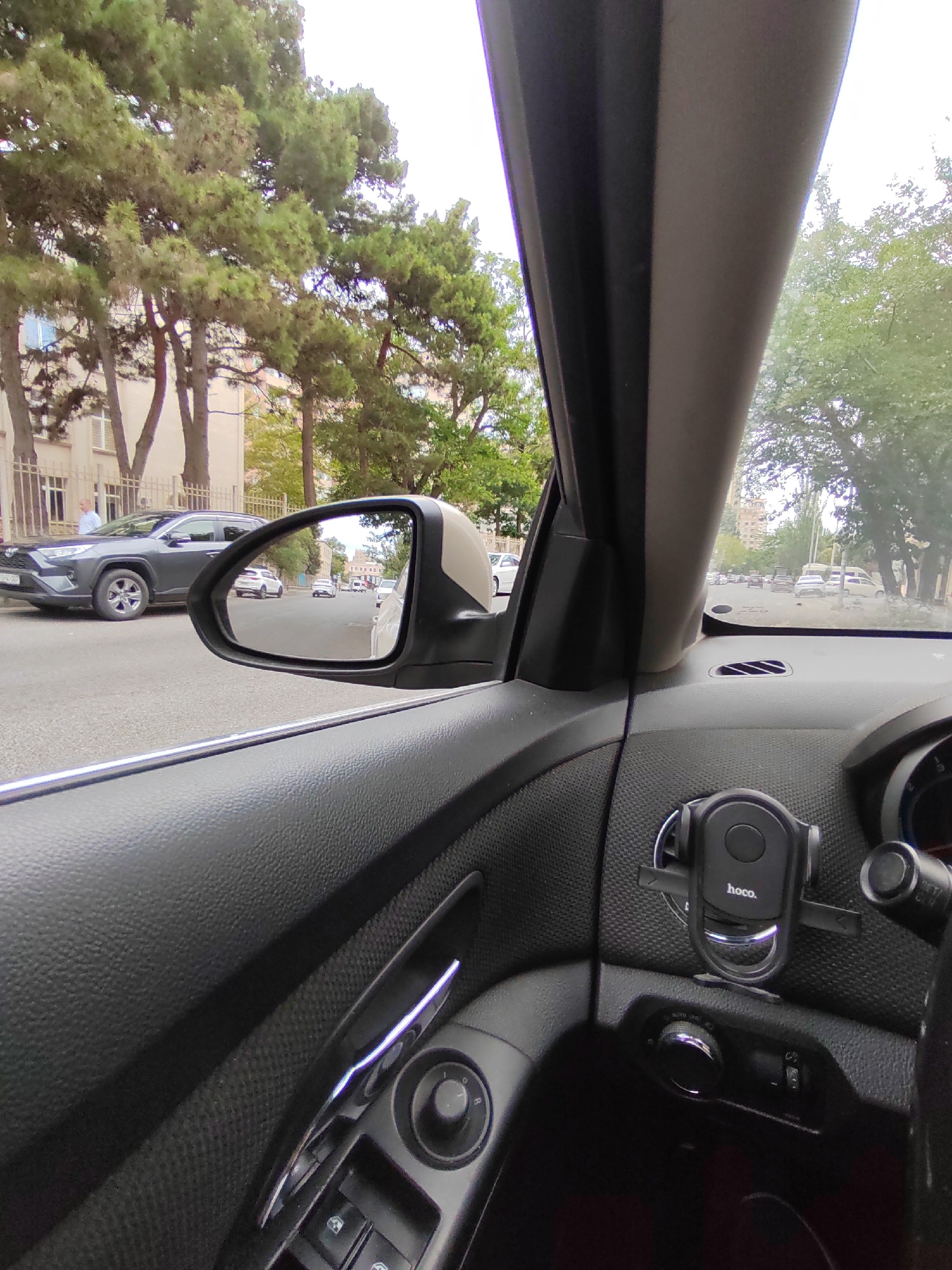 The image shows a view from inside a car, looking out through the windshield and side window, with a side mirror reflecting the road. The car is on a street in Baku, Azerbaijan, as indicated by the architectural style of the buildings and the presence of trees commonly found in the city. In the foreground, the interior of the car is visible, including the dashboard with a phone mount labeled "hoco.", a window control panel, and part of the steering wheel.
Outside the car, a dark grey SUV is parked on the left side of the road. Further down the street, several other cars are visible, moving in the same direction. The road is paved and appears to be a multi-lane street. Trees line the sides of the road, with lush green foliage. A building with a light-colored facade and balconies is visible behind the trees on the left. The sky is overcast, suggesting a cloudy day. The overall scene depicts a typical urban street view in Baku.
