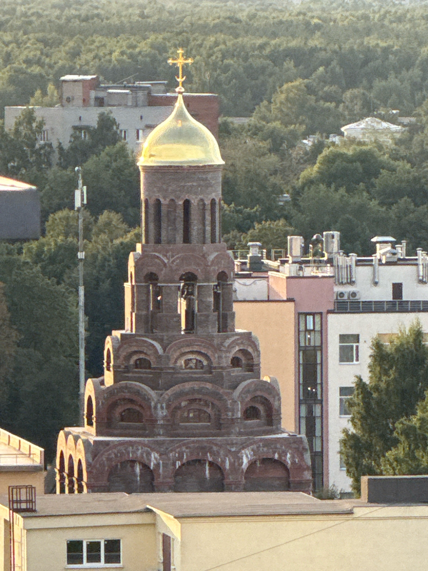 The image shows a view of an urban landscape with a prominent brick bell tower topped with a golden dome and cross. The bell tower is intricately designed with multiple tiers, each featuring arched openings and decorative brickwork. In the background, various buildings of different sizes and architectural styles are visible, including a taller, light-colored building with numerous windows and rooftop mechanical units. Green trees densely populate the area surrounding the buildings, suggesting a relatively green city or the presence of a park. A communication tower with antennas is also visible to the left of the bell tower. The lighting indicates it is daytime, likely late afternoon, with the sun casting shadows and highlighting the textures of the structures. The overall scene depicts a cityscape with a distinct religious architectural element.