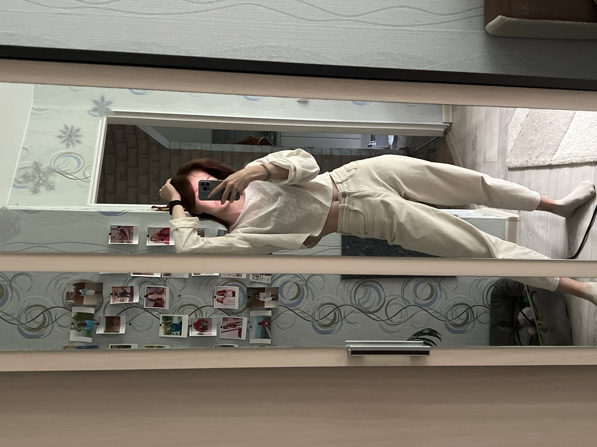 An indoor mirror selfie captures a young female subject lying horizontally on a surface, likely a bed or sofa, within a residence in Chaikovsky, Russia. She has light brown or reddish hair and is seen holding a dark-colored smartphone, which obscures her face, in her left hand. A black wristband is visible on her left wrist. She is dressed casually in a light-colored, long-sleeved top, possibly featuring eyelet or embroidered details, that exposes a small portion of her midriff. She also wears light beige or cream-colored pants and white socks.
The mirror's reflection reveals elements of the room. The wall behind her is adorned with distinctive wallpaper featuring blue, grey, and brown swirling patterns, along with several snowflake decals. Below the main reflection, a series of small, possibly instant-print photographs are taped to the wall or mirror frame. These pictures appear to depict people and various scenes, arranged along a wavy dark line. Other visible elements include a light-colored rug on the floor and a glimpse of a houseplant in the lower right area of the reflection. The overall lighting suggests it is daytime. There is no discernible text visible in the image. The scene conveys a casual and personal moment.
