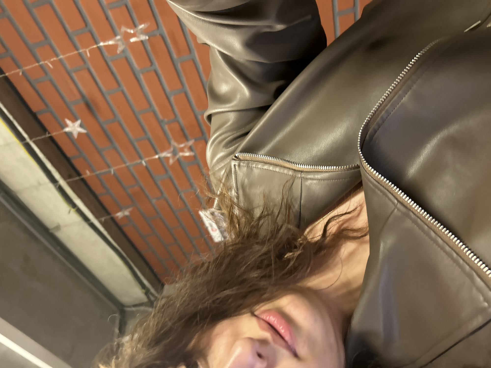 The image is a close-up, slightly off-center, low-angle shot of a person wearing a leather jacket. The jacket is partially zipped, revealing some clothing underneath. The person has curly hair and their nose and lips are visible. The background features a brick ceiling with decorative star-shaped lights. The location appears to be indoors, possibly a shop or an establishment with a brick ceiling and decorative lighting. There is no clear indication that this is in Tbilisi, Georgia.