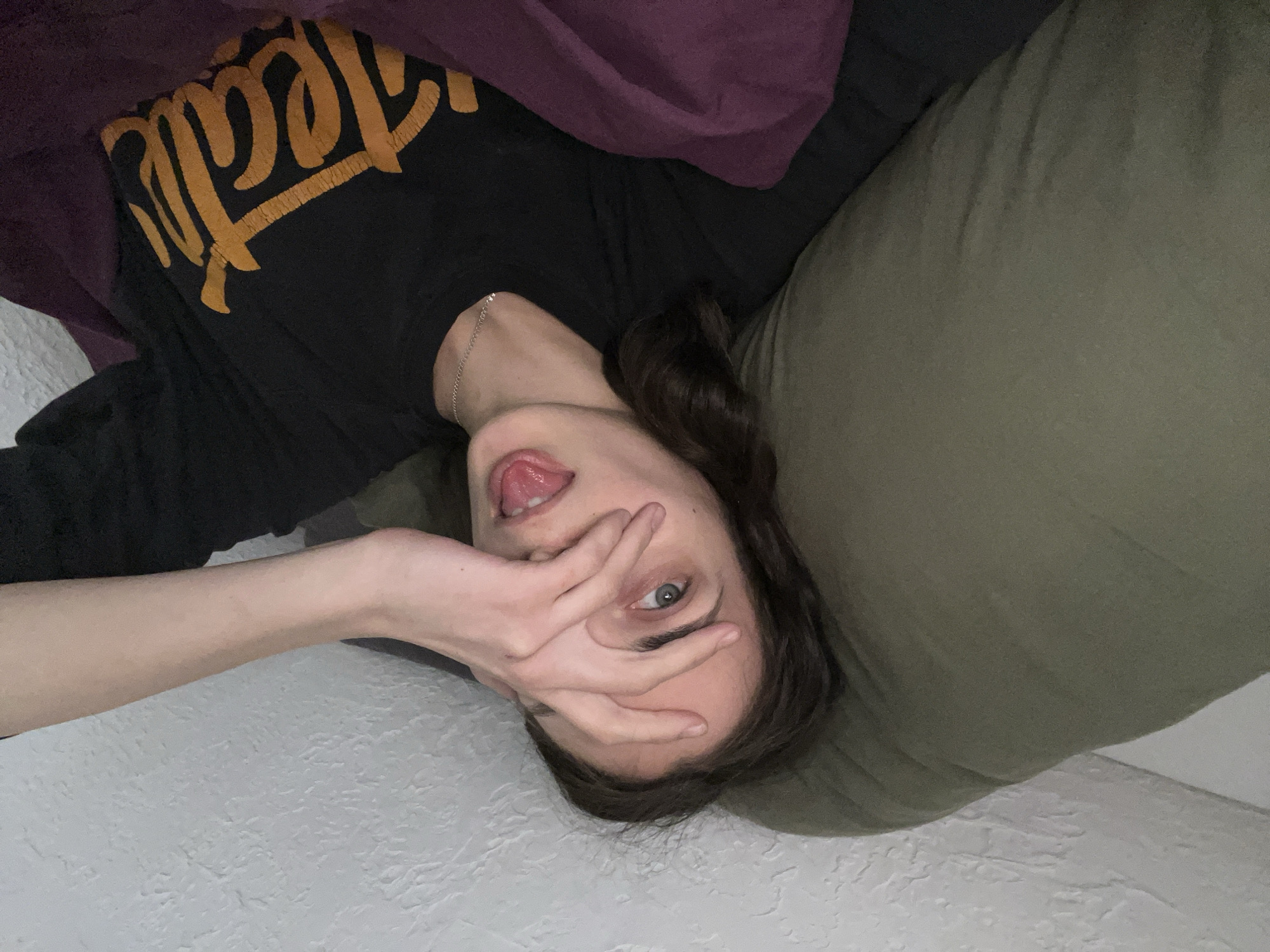 A person is lying down, partially covered by a green pillow and a purple blanket. The person is wearing a black shirt with yellow lettering, sticking their tongue out, and covering their face with their hand. The background consists of a white, textured surface. There is no information available to determine the location in Krasnoyarsk, Russia.