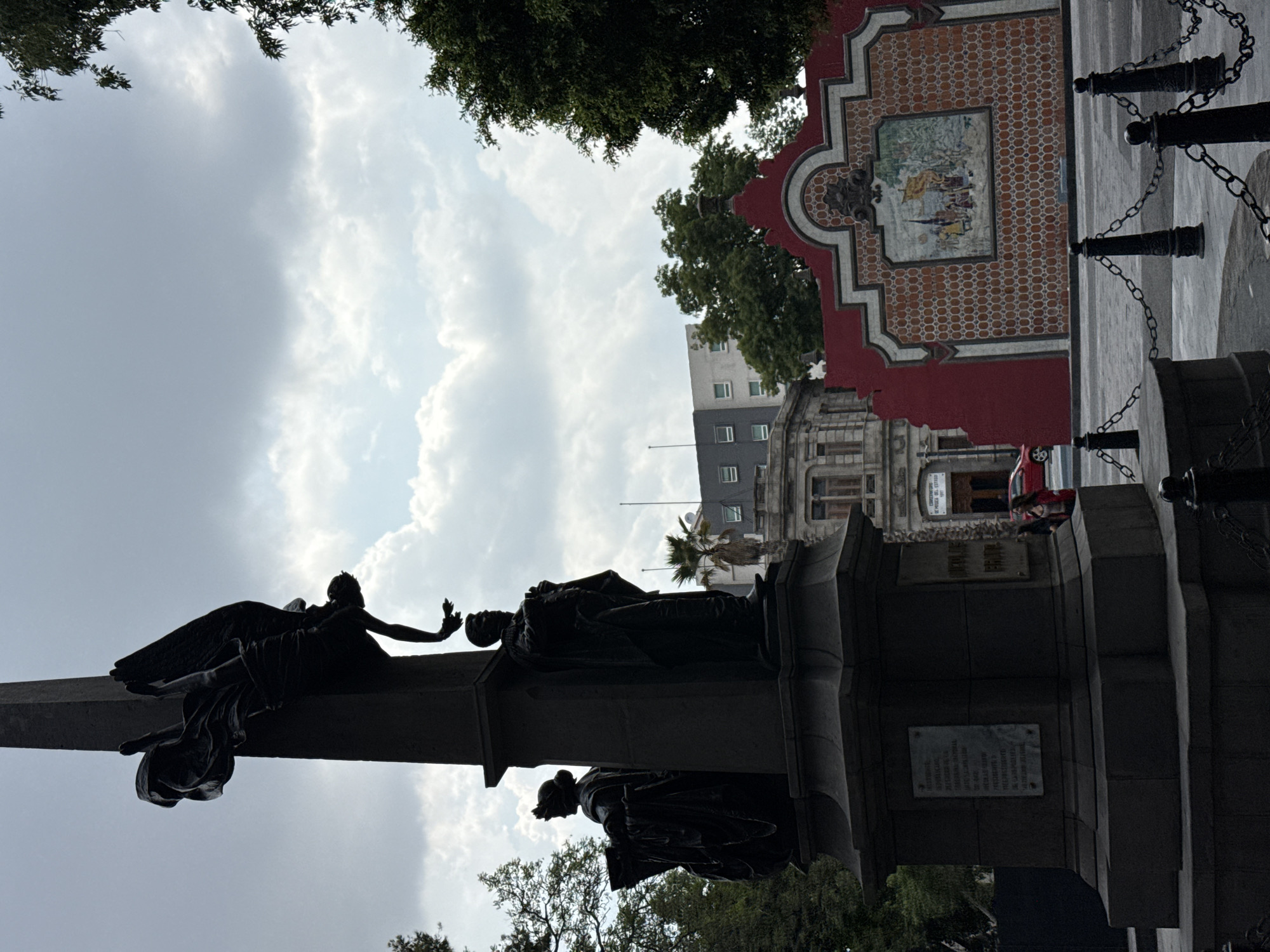 The image shows a monument in Puebla, Mexico, likely the Monument to the Founders of Puebla located in the Plazuela de los Fundadores. The monument features a column topped with a statue of an angel crowning a figure. Below, there are other statues and inscribed plaques. The monument is surrounded by trees and other buildings, including a red building with decorative tiles. The sky is cloudy.