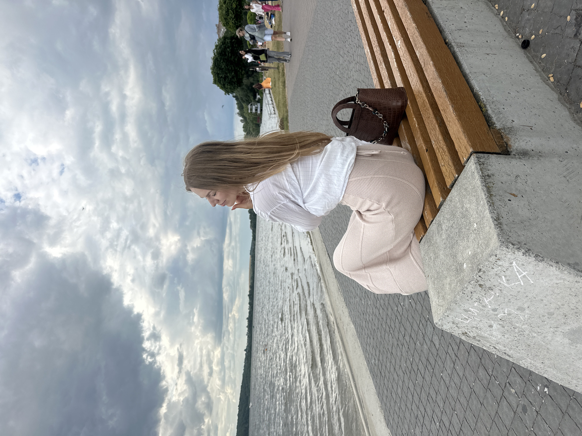 A young woman with long blonde hair, wearing a white shirt and a light pink skirt, sits on a wooden bench facing a lake. She is looking out at the water, with her eyes closed and her hand resting on her chin. A brown crocodile-embossed handbag is placed on the bench next to her. The bench is situated on a paved walkway by the lake. In the background, several people are walking along the path, and trees and buildings are visible on the far shore. The sky is cloudy and overcast. This scene is likely at Ternopil Lake in Ternopil, Ukraine.