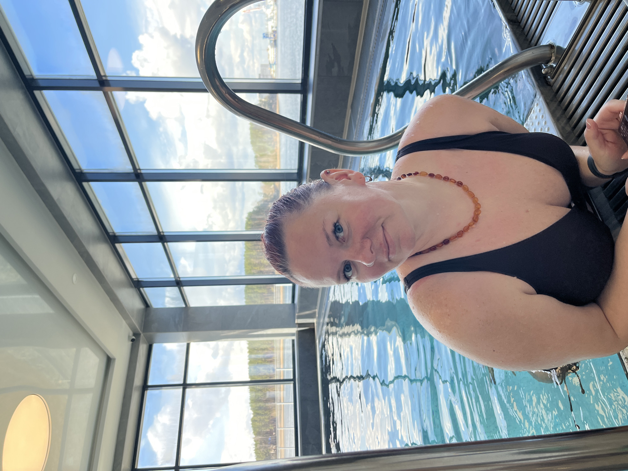 An image captured from a slightly low angle depicts a woman partially submerged in a swimming pool, looking directly at the camera with a gentle smile. She has wet, possibly reddish-tinted hair pulled back, striking blue eyes, and is wearing a black swimsuit and an amber-colored beaded necklace. One ear is visible with an earring, and a nose piercing can also be seen.
The setting is an indoor swimming pool facility, characterized by modern architecture featuring expansive, floor-to-ceiling windows that allow abundant natural light. Through these large windows, a clear blue sky with white clouds is visible, indicating a bright, sunny day. The windows frame a scenic view of a large body of water, likely a river or reservoir given the Chaikovsky, Russia context, with distant forested land. Some faint industrial elements, possibly cranes, can be discerned near the water's edge in the far distance.
The pool's surface is a light blue, reflecting the sky and showing ripples from the water. A sturdy metal handrail for pool entry/exit is prominent in the foreground and midground. The interior design includes sleek dark frames for the windows, concrete-like structural elements, and a white ceiling with a circular light fixture and a small security camera visible in one corner. The overall impression is one of relaxation in a bright, contemporary environment with a picturesque view. There is no visible text in the image.