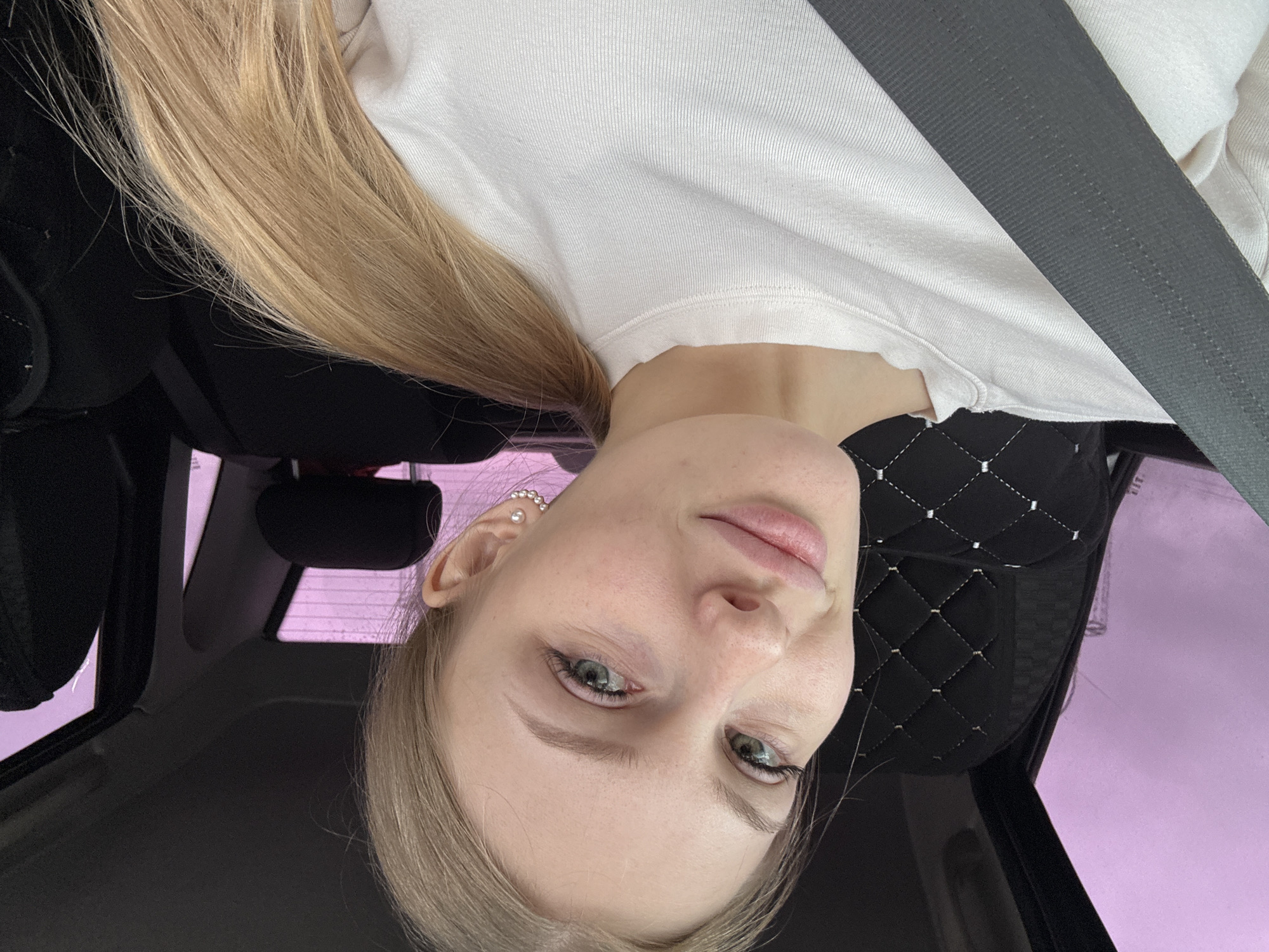 The image shows a young woman with blonde hair, fair skin, and green eyes wearing a white sweatshirt and a seatbelt in a car. She is looking directly at the camera with a slight smile. The car's interior is visible, including the black leather seats and headrest. The background shows a glimpse of the outside, with a purplish hue suggesting it might be dusk or dawn. There is no information to confirm the location in Perm, Russia.
