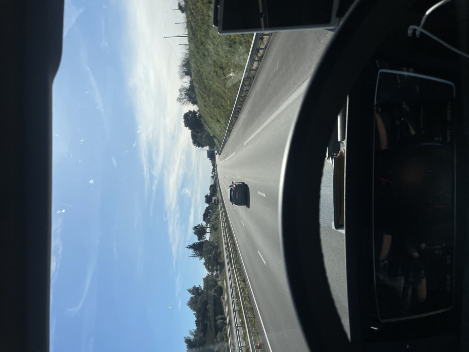 The image presents a view from inside a vehicle, likely a truck, on a multi-lane highway near Tavel, France, on a clear, sunny day. A dark steering wheel and a digital dashboard display, showing "14035 KM" among other indistinct information, are prominent in the lower foreground. Through the windshield, the paved highway extends into the distance, marked with white lines for at least two lanes in each direction. A single dark-colored passenger car, appearing to be a hatchback or small SUV, is positioned in the middle lane, moving away from the viewer. Metal guardrails line both sides of the roadway, separating it from grassy embankments adorned with scattered trees and shrubs. The sky is a bright blue with sparse, wispy white clouds, and direct sunlight causes glare on the windshield. Additional vehicles are faintly visible in the far distance on the highway, and utility poles stand on the right-side embankment further down the road.