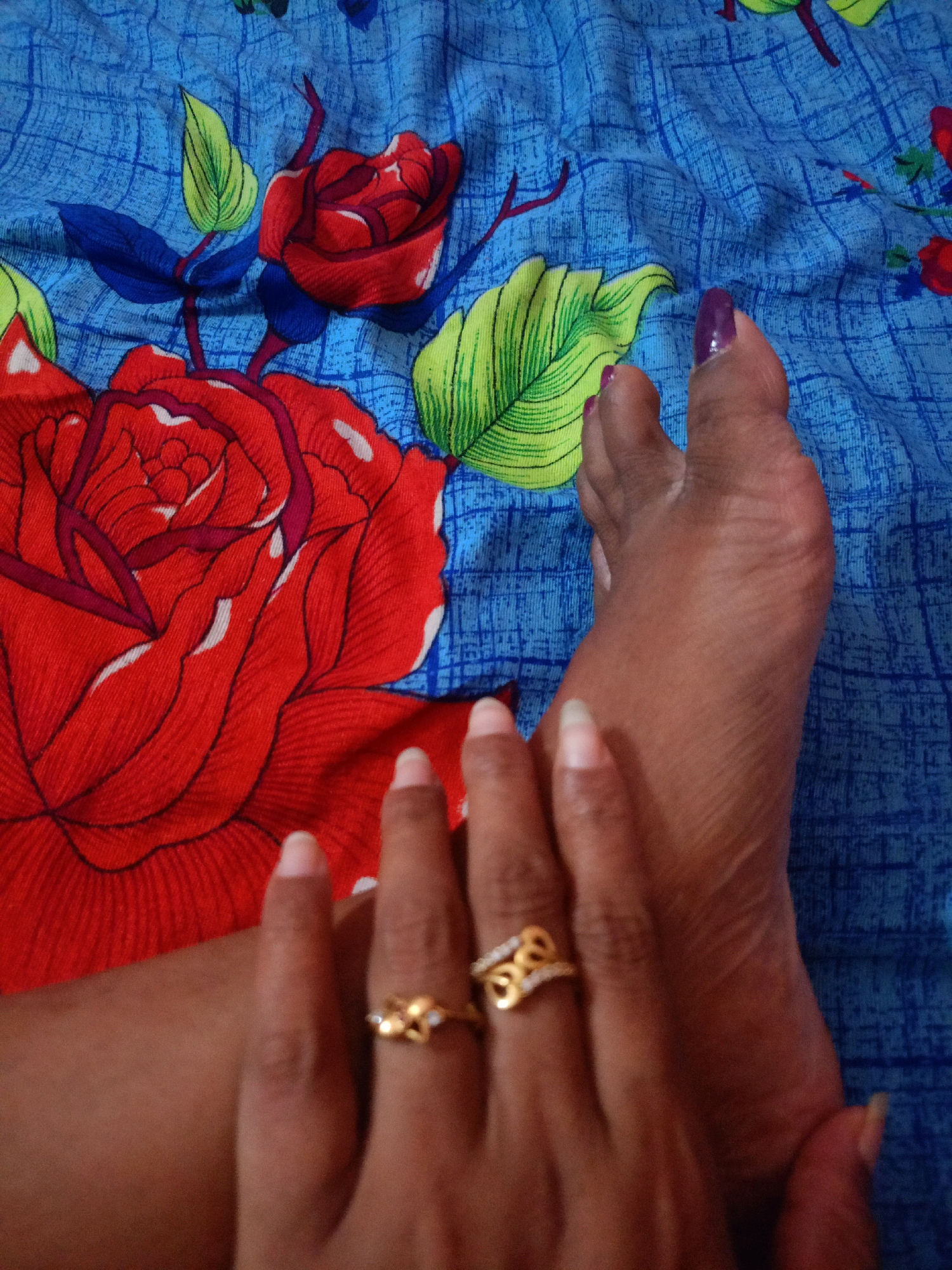 A close-up, overhead shot captures a person's feet and hands against a blue fabric background with a red rose pattern. The fabric exhibits wrinkles and folds, indicative of being a bedsheet or similar textile. The right foot, with toes pointed towards the upper left, has purple nail polish on the toenails. The skin on the foot shows texture and creases. The left hand rests on the foot, with fingers adorned with two gold rings. The ring on the index finger features small diamond-like accents in a floral pattern, while the ring on the middle finger is a band with an abstract design and diamond-like accents. The fingernails are unpolished. The overall scene appears to be an indoor setting, likely a bedroom, with no discernable geographical markers to pinpoint a location in Chenkavila, India.