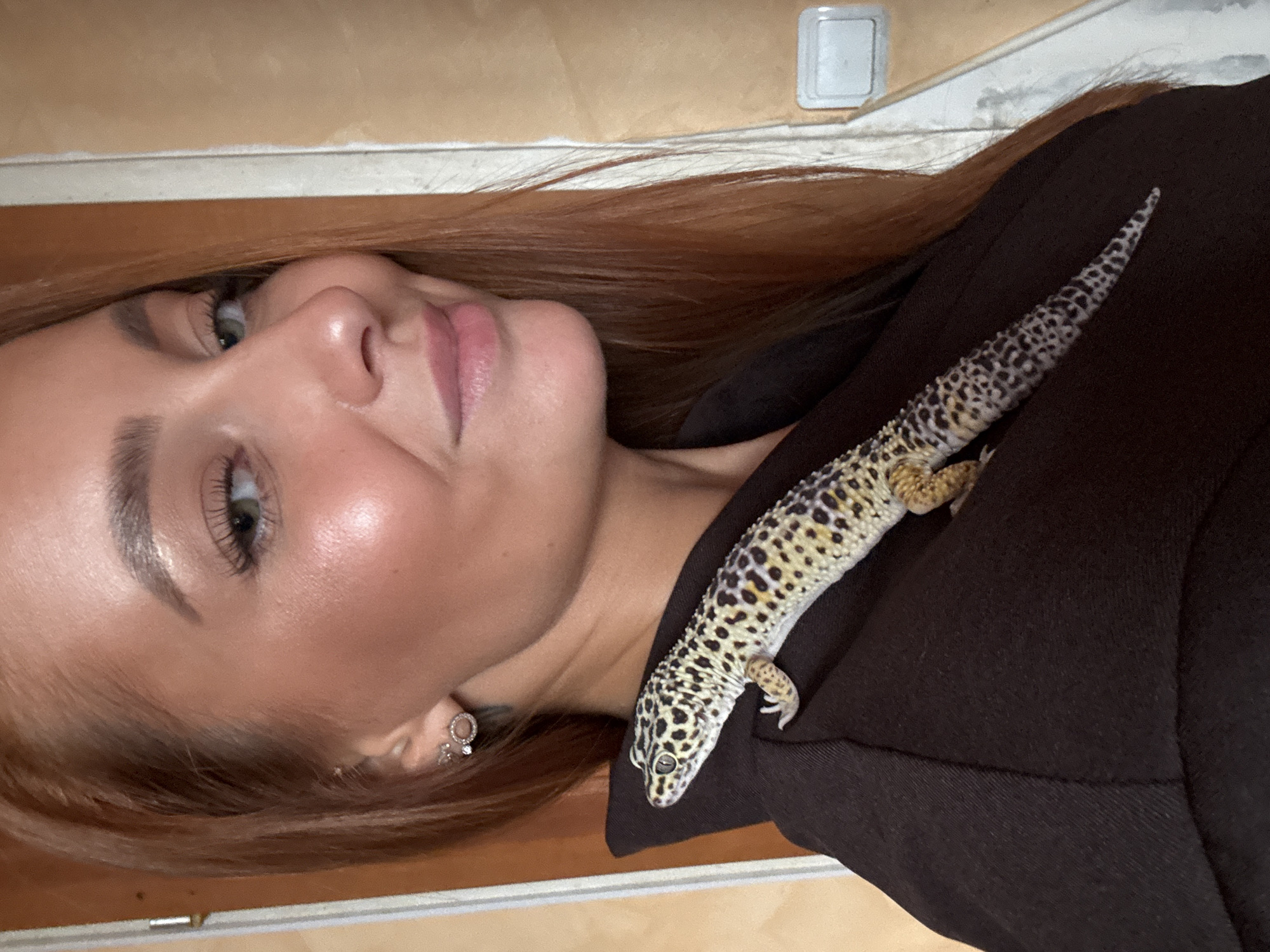 A woman with auburn hair is positioned close to the camera, her head tilted to the left. A leopard gecko rests on her right shoulder, its body stretched across her dark-colored garment. The woman's face is partially visible, showcasing her fair complexion, light green eyes, and pink lips. She is wearing earrings. In the background, there's a light switch on a wall. The location is unspecified, and there is no verifiable evidence to confirm the location as Chaikovsky, Russia.