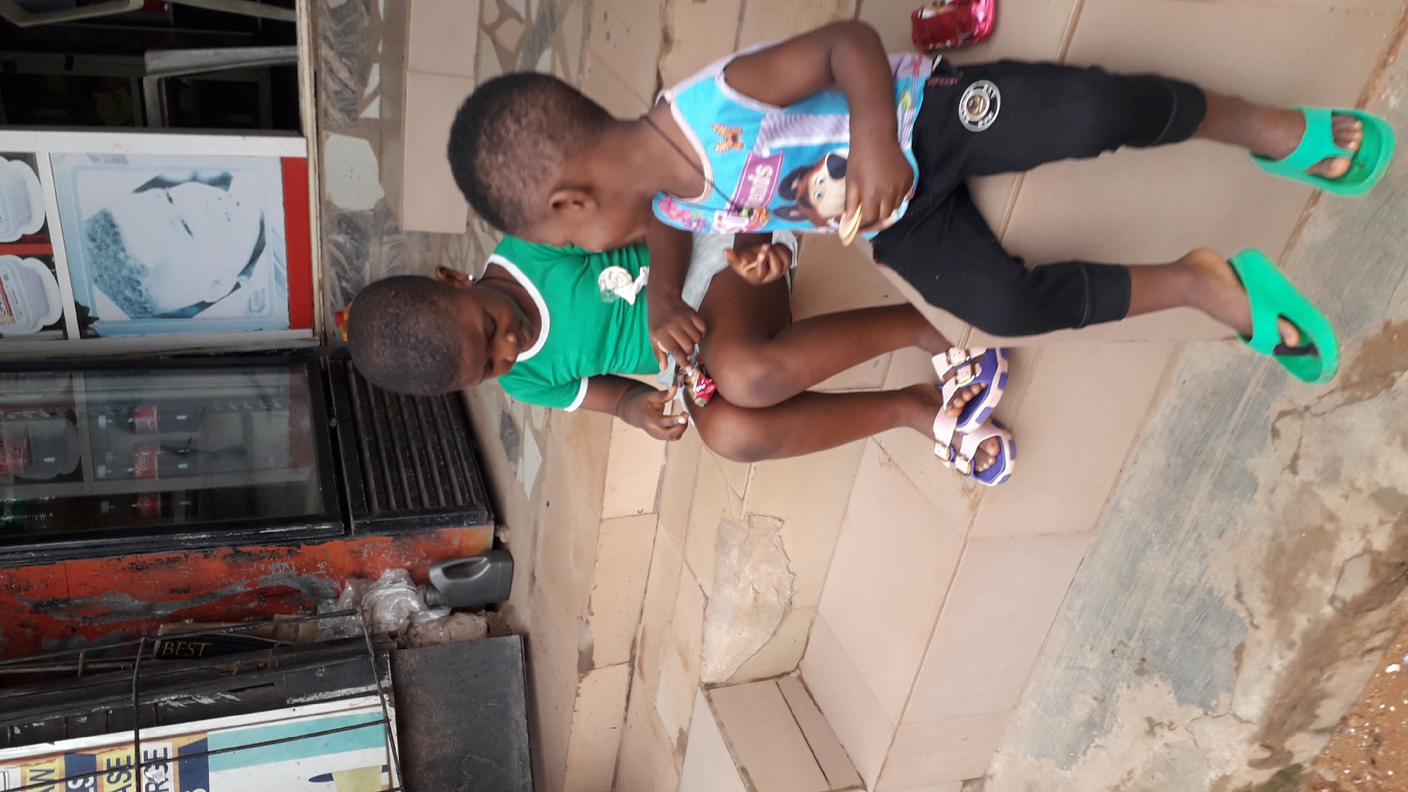 Two young children, a girl and a boy, are sitting on a set of tiled steps outside a building. The girl, on the left, is wearing a green t-shirt and light-colored shorts, with purple and white sandals. She is holding a wrapped candy in her hands and appears to be examining it. The boy, on the right, is wearing a sleeveless blue and white shirt with a character graphic and black pants, with green flip-flops. He is holding a piece of food, possibly a biscuit or cracker, and looking towards the girl. Behind them, a commercial refrigerator with glass doors displays bottled beverages. A poster with a black and white image of a child's face is affixed to the wall. The environment suggests a retail or commercial setting, possibly a small shop or kiosk in Nkpor, Nigeria, given the tiled steps and the presence of a commercial refrigerator. The steps themselves show some signs of wear and tear.