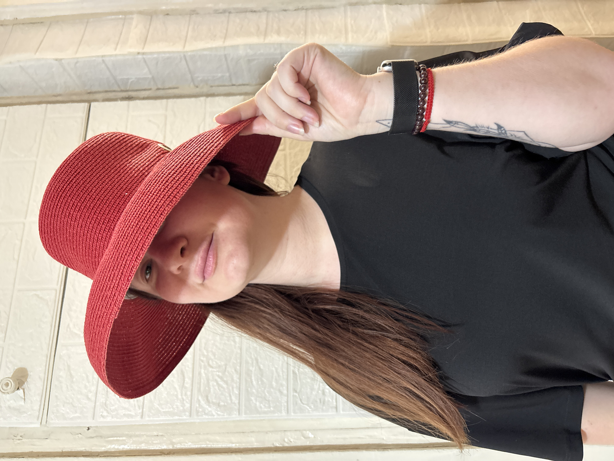 A young Caucasian woman stands in front of a white, textured wall, possibly in a residential setting in Sochi, Russia. She is wearing a black t-shirt and a wide-brimmed red straw hat. The hat casts a shadow over her eyes, but her mouth and chin are visible. She is holding the brim of the hat with her right hand. On her right wrist, she wears a black smartwatch and two bracelets, one red and one with dark beads. She has a tattoo on her forearm. The background is a plain white wall with a textured pattern.