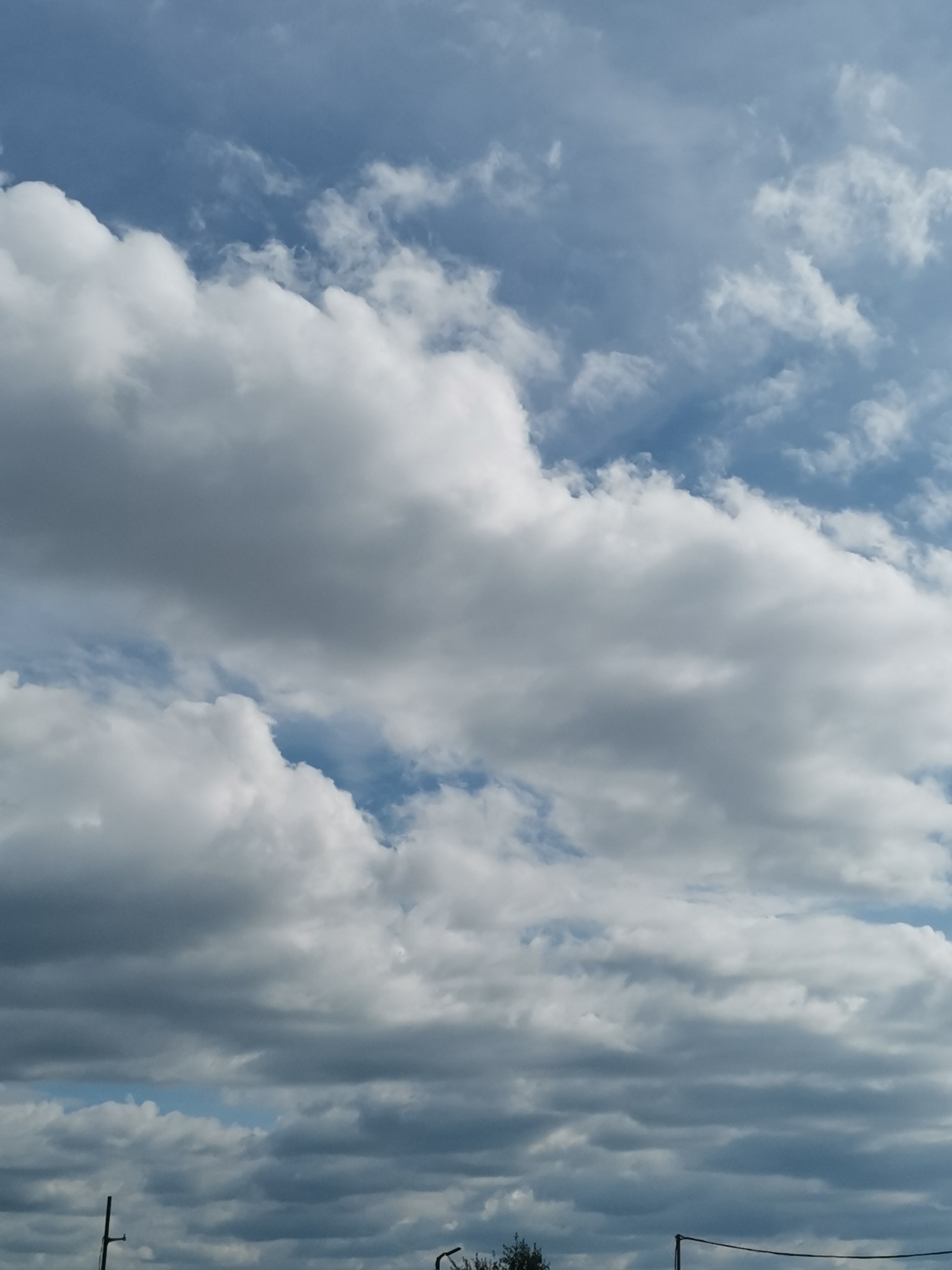 The image displays a cloudy sky. The clouds are varied in texture and density, ranging from wispy cirrus clouds to heavier cumulus formations. Some patches of blue sky are visible between the clouds. The lower portion of the image shows the top of a few indistinct structures, possibly electrical poles or antennas, and some foliage. The lighting suggests it is daytime. There are no identifiable people, specific landmarks, or any activity taking place that would indicate a particular location within Sofia, Bulgaria.