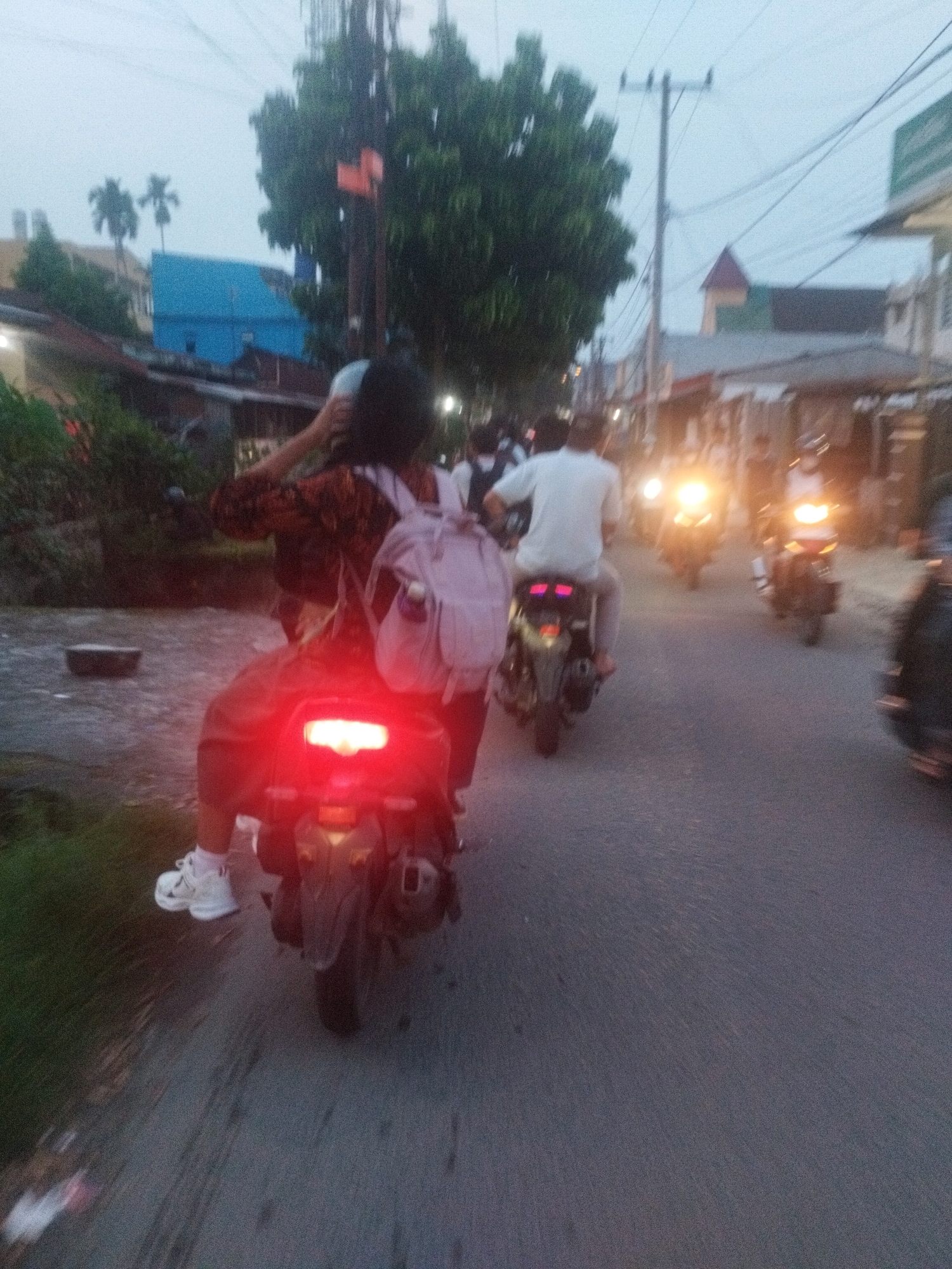 A group of motorcyclists are captured from behind in a street scene in the city of Medan, Indonesia, during dusk or early evening. The primary focus is on two individuals riding a scooter, with the passenger in the foreground wearing a patterned shirt and a light pink backpack. The scooter's red taillight is illuminated. Another motorcyclist, wearing a white shirt, is visible ahead of them, also riding a scooter. Additional blurred figures and motorbikes are present further down the street. The environment appears to be a roadside with buildings and vegetation on the left side, and streetlights or vehicle lights indicating urban activity. The sky is overcast, contributing to the low-light conditions.