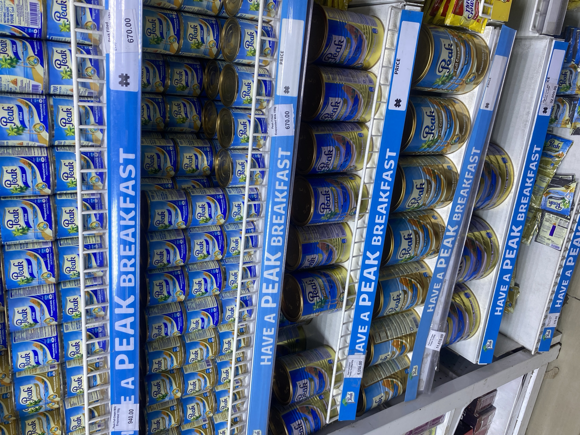 The image shows a shelf in a store, likely in Lagos, Nigeria, stocked with cans of Peak brand milk products. The cans are neatly arranged in rows and columns. Blue shelf-edge strips with white text reading "HAVE A PEAK BREAKFAST" are visible along the shelves, as well as signs with pricing and product information. There are multiple varieties of Peak milk, including Peak Filled Evaporated Milk, Peak Full Cream Milk. The prices displayed are 670.00, 940.00, 5,550.00, 11,570.00, 170.00, and 32,000.00.