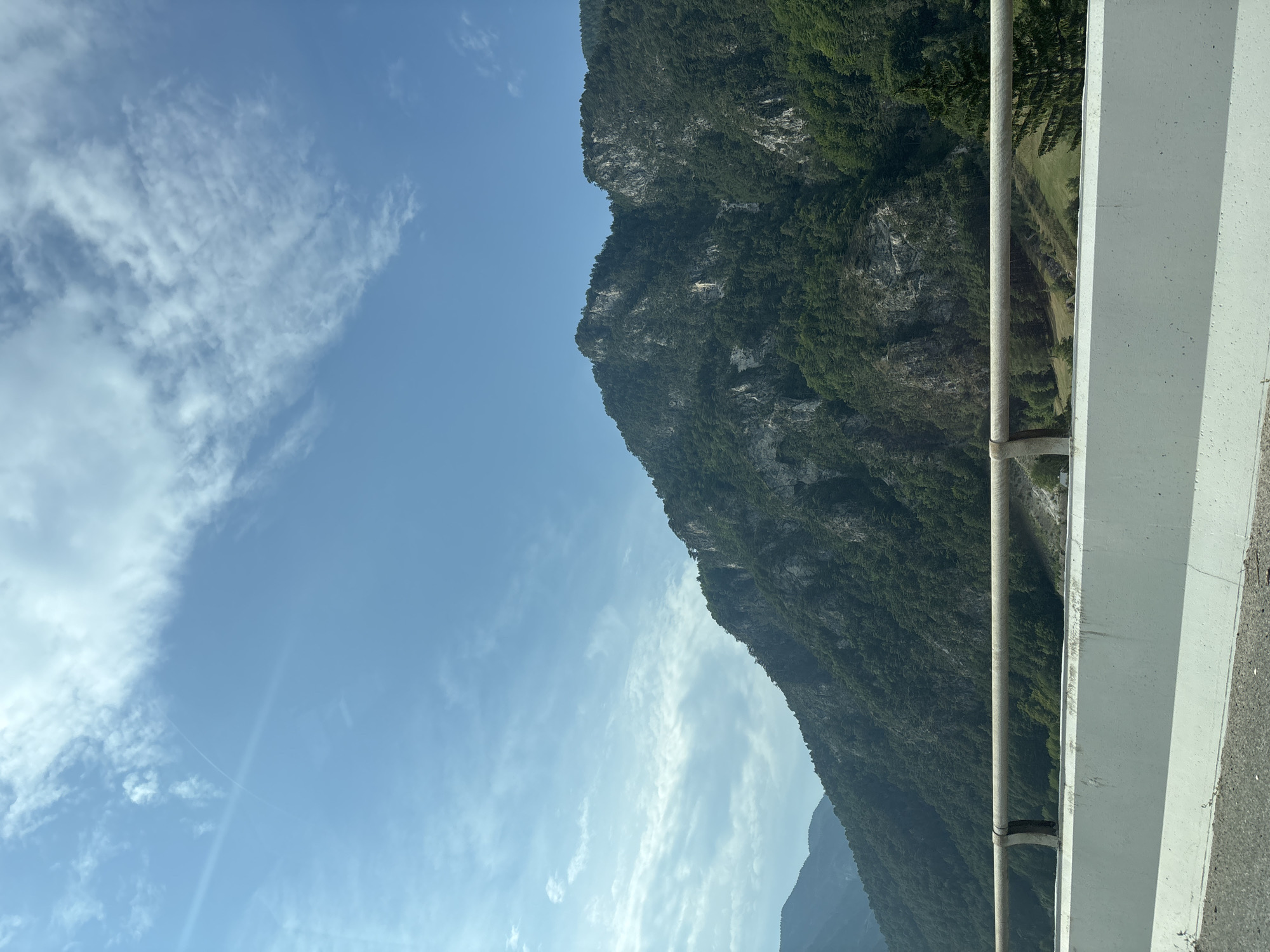 This is a photograph of a mountainous landscape under a bright, blue sky with scattered white clouds. The mountains are covered in dense green trees, with visible rocky outcrops and sheer cliffs. In the foreground, there is a white concrete barrier and a metal guardrail, suggesting a roadside or overlook. The scene is devoid of any people or animals. The lighting indicates it is daytime, likely with the sun positioned to the left, creating some lens flare and highlighting the textures of the mountains. The environment is natural and appears to be a scenic viewpoint in a mountainous region of Italy, possibly in the municipality of Malborghetto-Valbruna, known for its alpine scenery.
