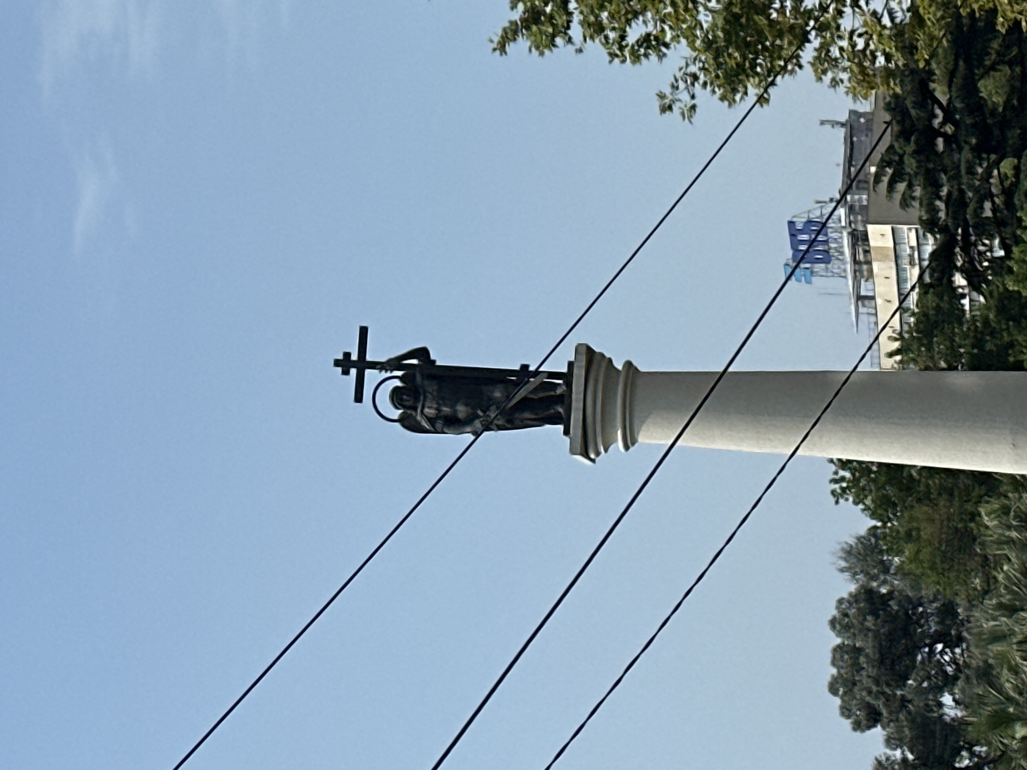 An outdoor, medium-shot photograph taken from a low angle shows a statue of an angel holding a cross atop a white column, situated in an urban setting. The sky is a clear, pale blue. Several dark, thin wires crisscross the frame from left to right. The angel statue is dark in color, possibly bronze, and has a halo. It stands on a white pedestal which is part of a taller, fluted white column. In the background, behind the column and to the right, a modern building with a prominent blue sign reading "2GIS" is visible, partially obscured by green foliage from trees. The trees in the foreground and mid-ground are lush and green, with some appearing to be palm trees. The overall impression is a clear day in a city, with a prominent monument set against a backdrop of modern infrastructure and vegetation.