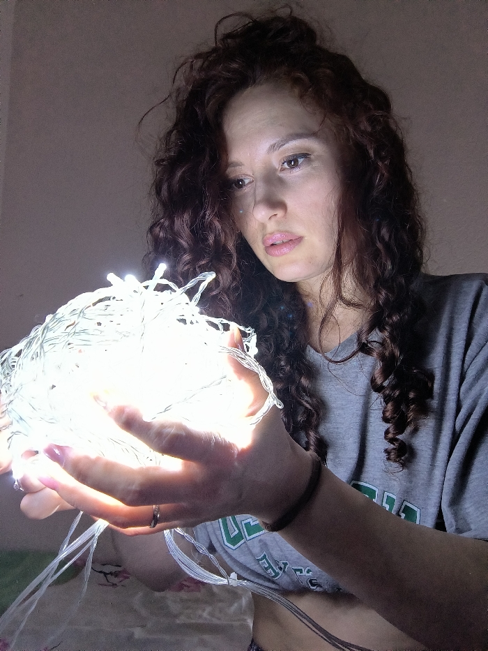 The image depicts a female individual, appearing to be in her 20s or 30s, situated in what is reported as Saint Petersburg, Russia. She is shown from the chest up, holding a spherical bundle of brightly illuminated white LED string lights in both hands, cupping them from below. Her long, curly reddish-brown hair cascades around her shoulders. Her face is directed downwards, with her gaze fixed intently on the glowing lights, which serve as the primary light source. The strong light from the LEDs creates significant glare and reflections on her fair skin and highlights her facial features. She wears a grey short-sleeved t-shirt with a partial green and white graphic visible on the lower right side, revealing a segment of her abdomen. A dark wristband is present on her right wrist, and a silver-toned ring is worn on her left ring finger. Her fingernails exhibit a light pink polish. The background consists of a uniformly light-colored, out-of-focus wall, dimly lit by the glow of the string lights.