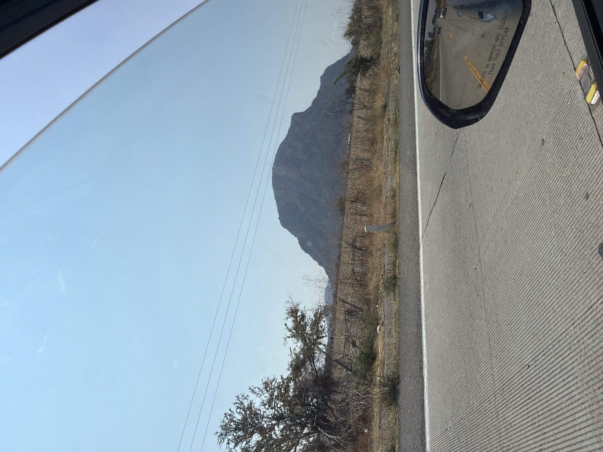 The image, taken from inside a moving vehicle, depicts a view of the landscape outside the passenger window. A large mountain looms in the distance against a light blue sky, with power lines running across the view. In the foreground, there are dry shrubs and trees along the side of the road. The road itself, appearing gray and textured, is visible with a white dividing line. The car's side mirror is present in the upper right corner, reflecting the road and another vehicle. The general scenery suggests a rural or semi-arid environment. There are no indications of Jantetelco or any other specific location or city. The image depicts no people.