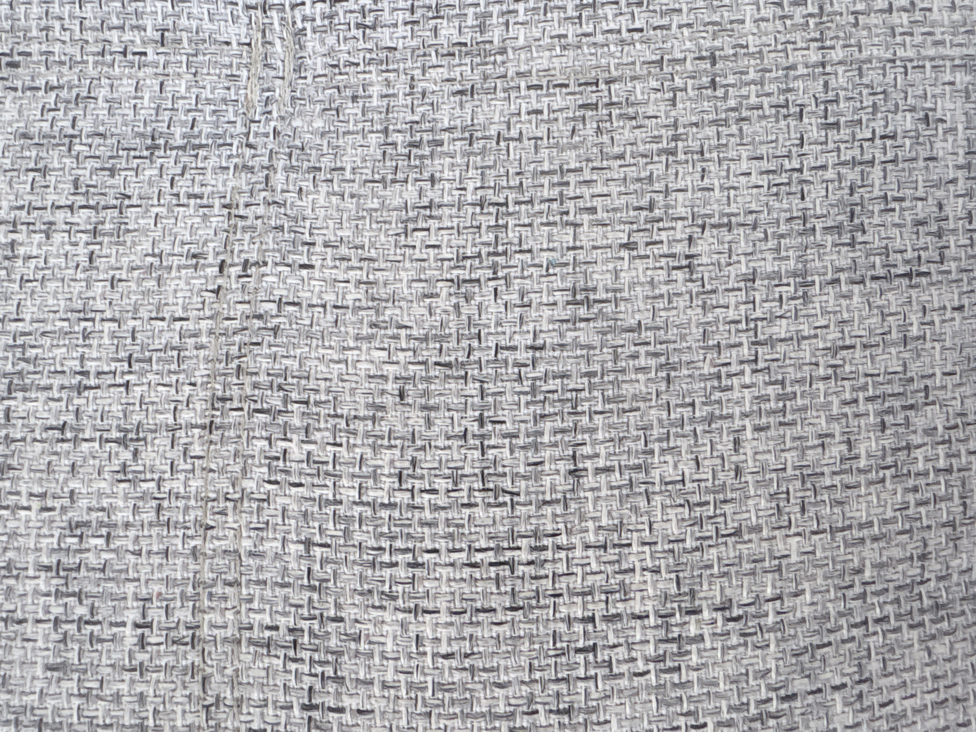 The image is a close-up, overhead shot of a textured grey fabric. The fabric appears to be woven with a tight, interlocking pattern of threads. Some threads are lighter grey, while others are darker grey, creating a subtle visual variation. The texture is coarse and appears to be made of natural fibers, possibly linen or a similar material. There is a slight sheen to the fabric in some areas, suggesting it might be a blend. A thin, lighter-colored thread is visible horizontally across the middle right portion of the image, possibly a snag or a loose end. The image is devoid of any context related to the city of Vyazniki, Russia, or any other specific location. It exclusively focuses on the textile's surface.
