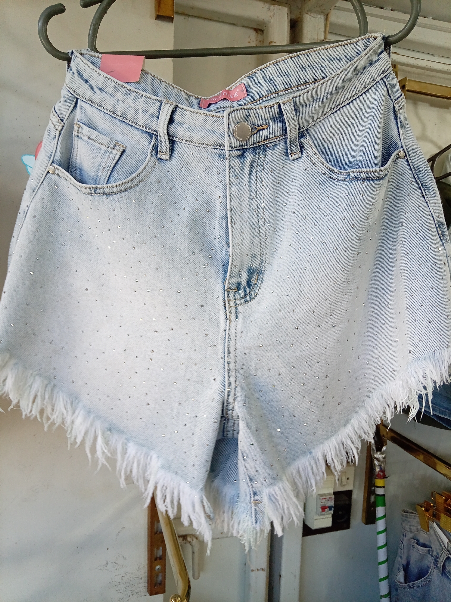 The image shows a pair of light-wash denim shorts with a frayed hem, adorned with small, scattered rhinestones. The shorts are hanging on a clothes hanger against a neutral background. The shorts appear to be displayed in a retail setting, as suggested by the presence of other clothing items and display racks in the background. The overall impression is of a fashion item being presented for sale. The image provides no information about the city of Donetsk, Ukraine, or any specific environmental details that would place it in that location.
