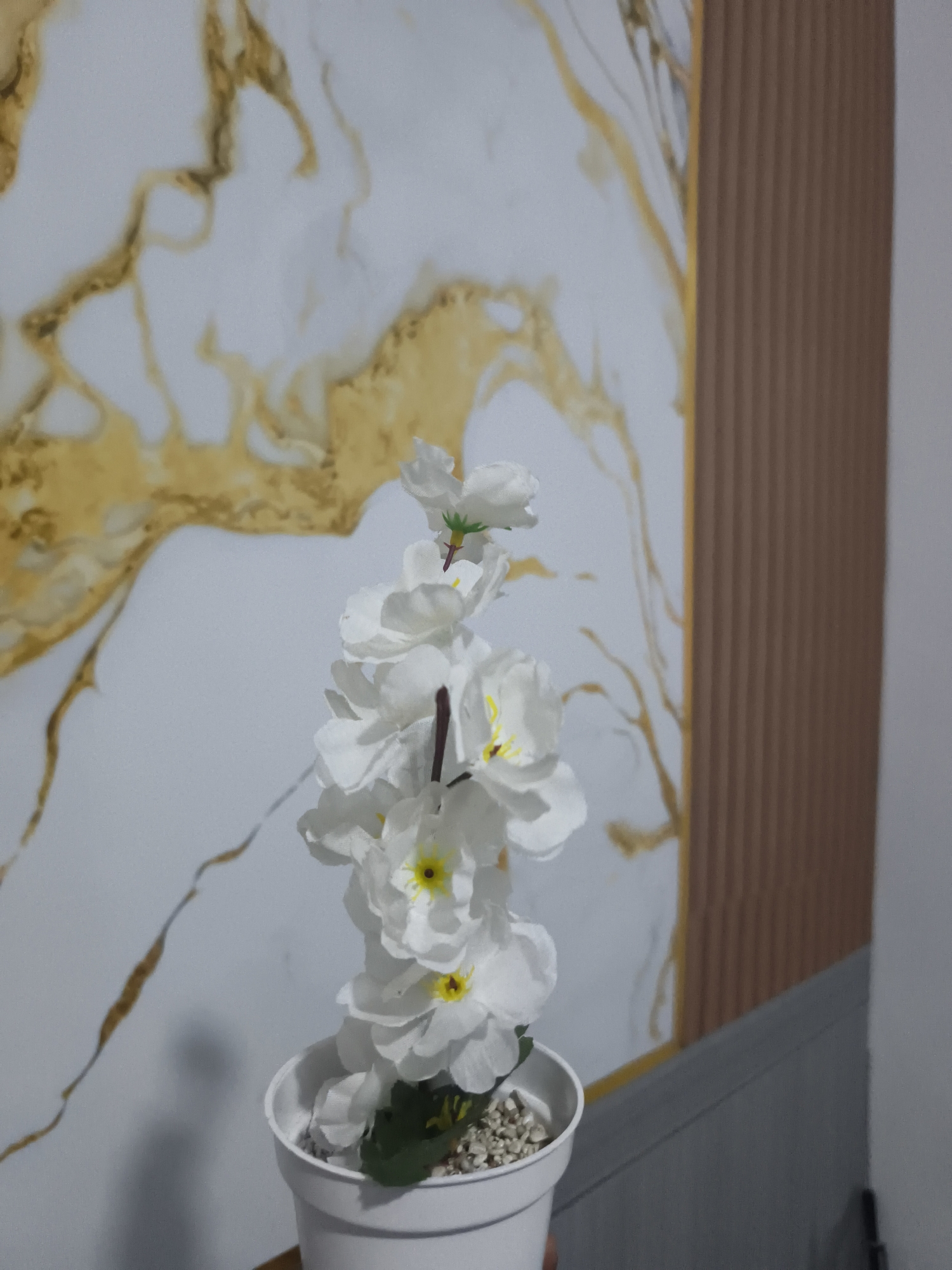 A close-up shot focuses on a white artificial flower arrangement in a white pot. The flower stalk, adorned with multiple white blooms with yellow centers and green leaves, extends upwards. The background features a textured wall with a marble-like pattern in white and gold, and a vertical brown ribbed panel on the right side. The scene is set indoors.