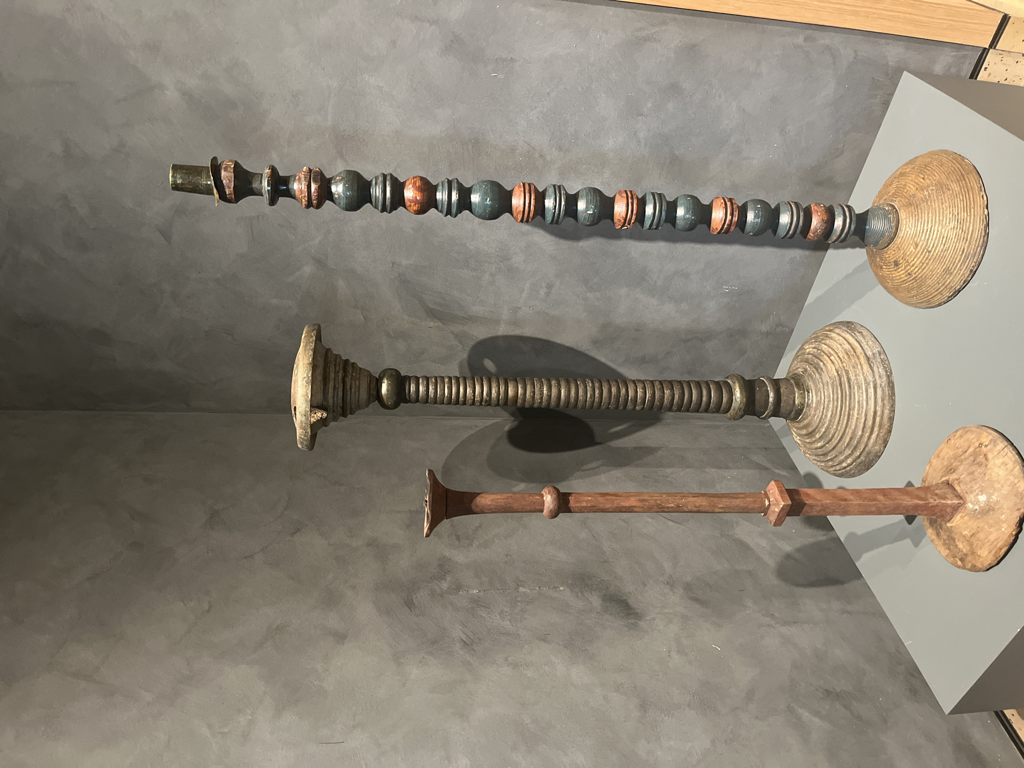 The image displays three decorative antique objects mounted horizontally on a gray wall in an indoor setting, location Unknown, Unknown. The objects resemble ornate candle holders or ceremonial staffs.
The uppermost object features alternating dark green and light brown spherical beads along its length, terminating in a circular base. The middle object is cylindrical with a ribbed texture, flaring out at both ends. The bottom object is a straight, dark brown rod with a small flare at one end and a larger, circular base at the other. The objects are mounted on a gray display platform affixed to the wall.