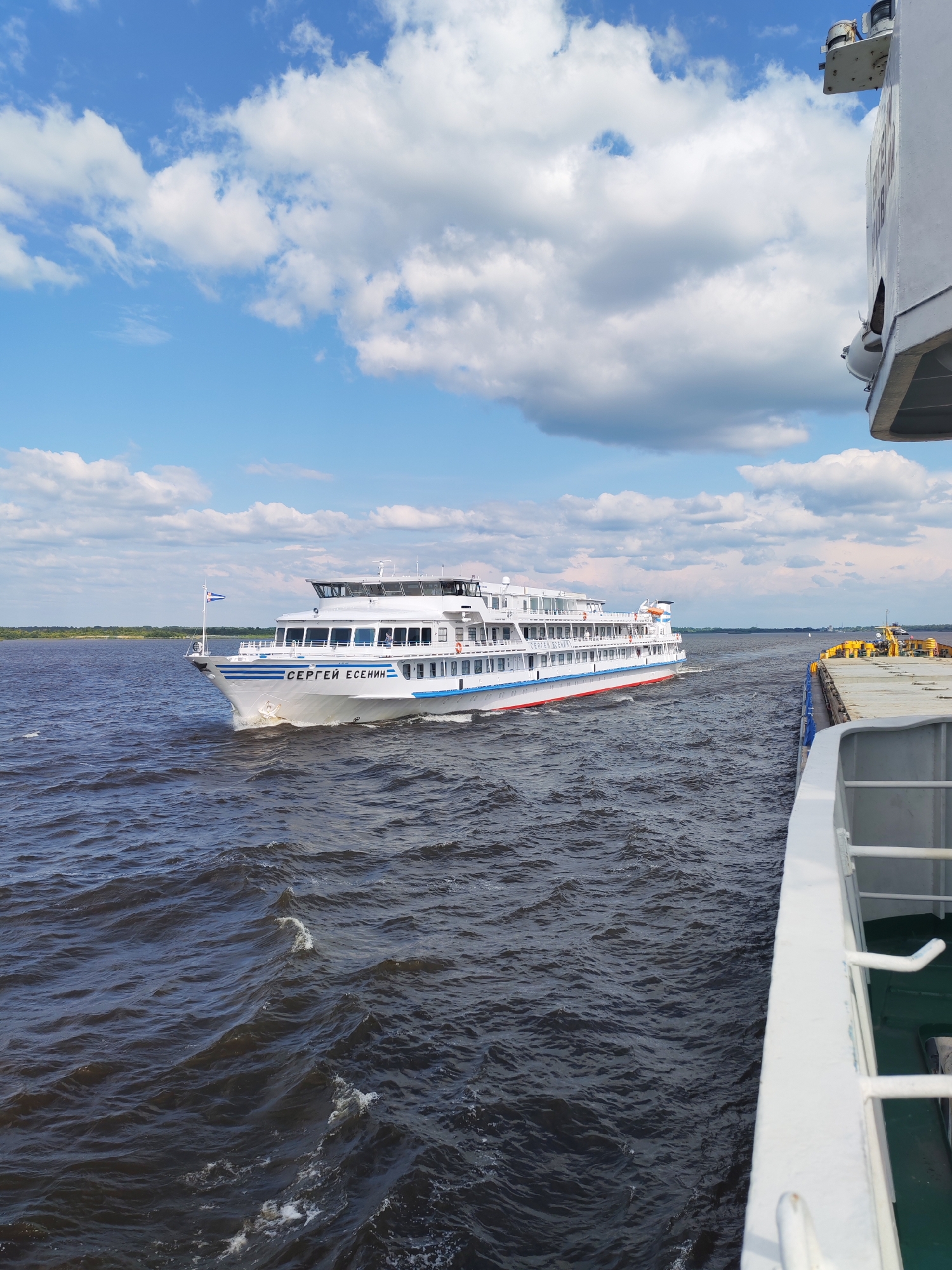 The image depicts a bright, sunny day on a body of water, possibly a river, with a blue sky filled with white, fluffy clouds. In the middle ground, a white passenger ship named "СЕРГЕЙ ЕСЕНИН" (Sergei Yesenin) is navigating the water. The ship has multiple decks, white railings, and blue and red stripes along its hull. A flag with a blue and white pattern is flying from the bow. In the foreground, a portion of another vessel is visible on the right, appearing to be a cargo ship with yellow structures on its deck. The water is dark and choppy, with visible waves. The background shows a distant, tree-lined shore under the bright sky. The location is specified as the city of Krokhino, Russia, though this detail cannot be independently verified from the visual content of the image alone. The scene suggests movement and travel on a waterway.
