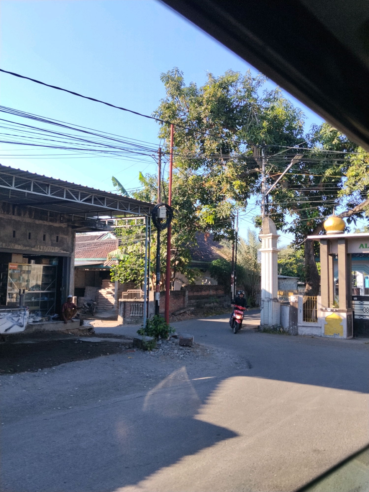 A motorcycle is seen riding down a paved road in Selong, Indonesia. The road is lined with buildings and trees. The sky is clear and blue. A person is sitting on the ground in front of a building. The building appears to be a shop, with glass display cases. There are several power lines overhead. In the background, a structure with a dome is visible.