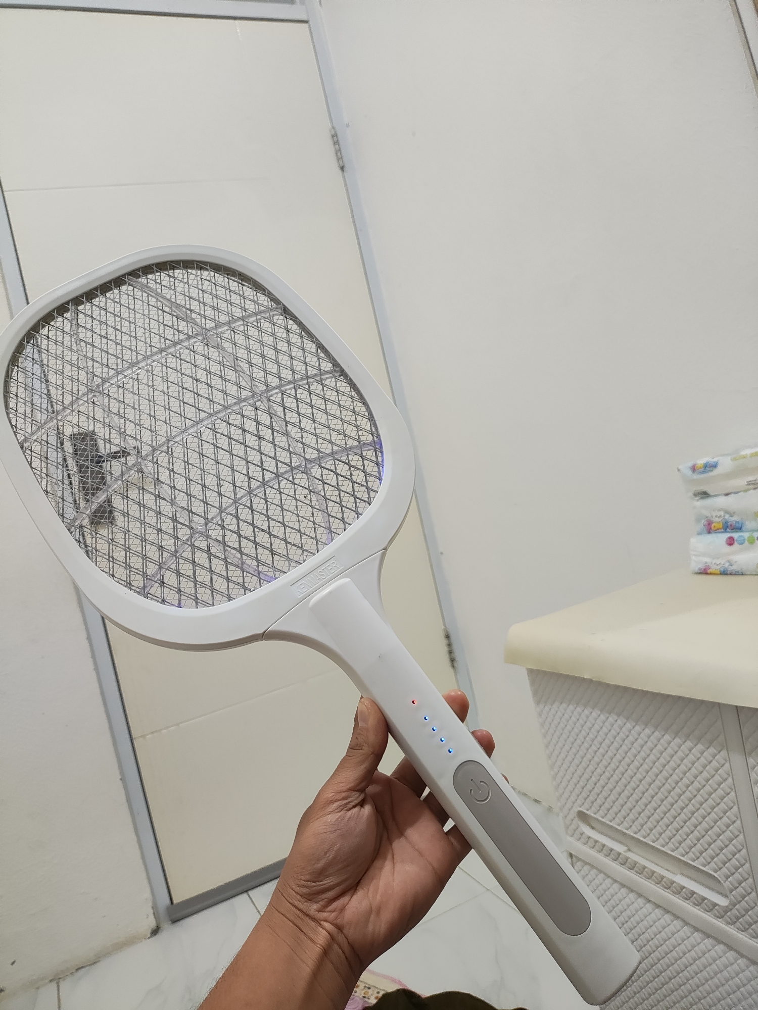 A person's hand holds a white electric mosquito swatter in the city of Wanajaya, Indonesia. The swatter, shaped like a tennis racket, has a metal grid with a diamond pattern. A shadowy insect is visible on the grid. The handle of the swatter features several indicator lights and a power button. In the background, a stack of diapers rests on a light-colored surface. A door and a plain white wall complete the indoor scene.