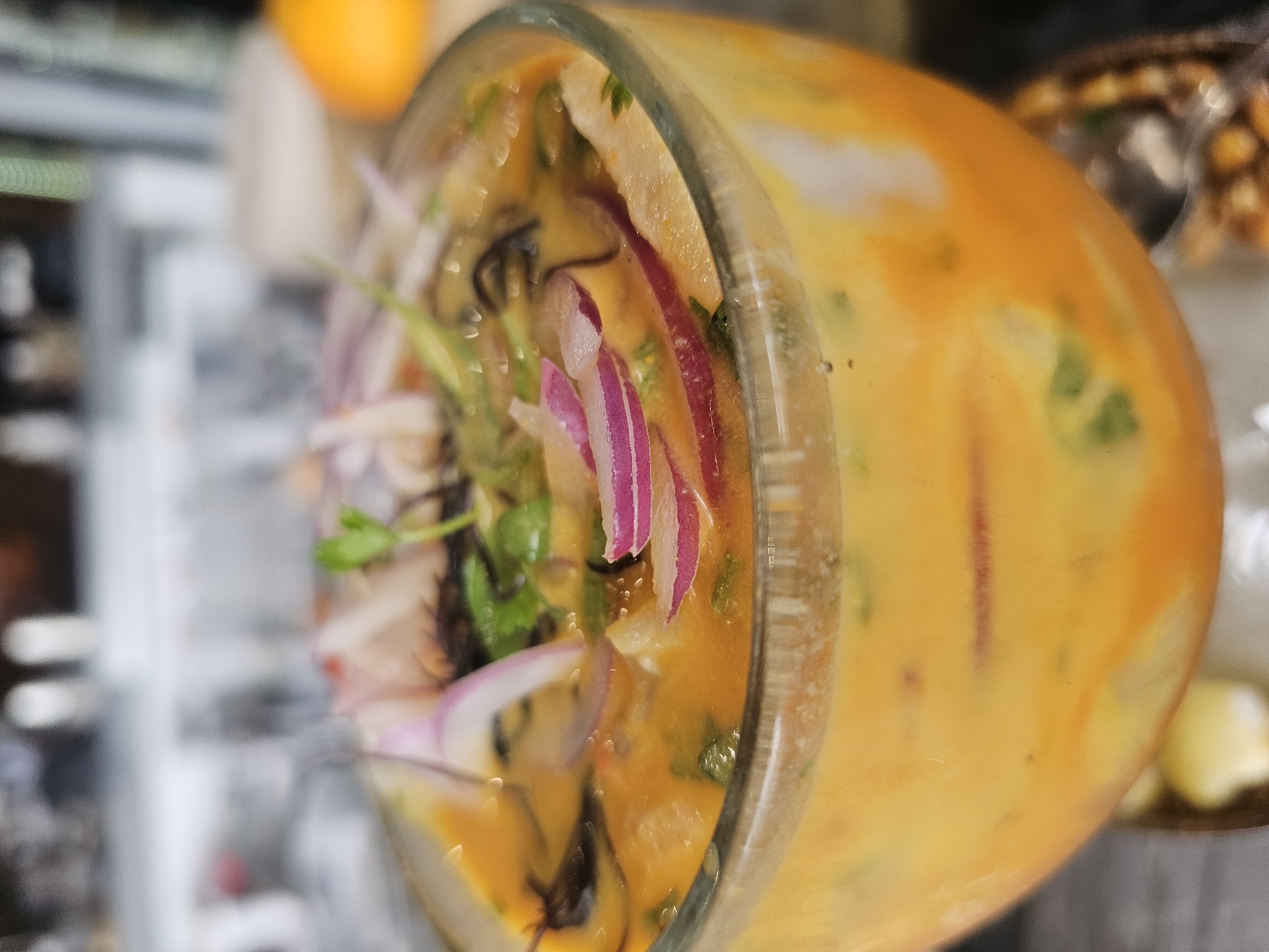 A close-up, vertically oriented image displays a clear glass filled with a vibrant orange-yellow liquid, strongly suggestive of a Peruvian dish like ceviche or leche de tigre. The glass is positioned horizontally across the frame, with its contents sharply in focus. Within the glass, thinly sliced red onions with their distinct purple and white layers are prominently visible, interspersed with fresh green cilantro leaves. Lighter-colored chunks, likely fish or seafood, are submerged in the opaque, richly colored liquid. Darker, thin, curly strands, possibly seaweed or a dark herb, add textural variation.
The background is softly blurred, providing context without distracting from the main subject. To the upper left, a bright orange fruit, possibly a mandarin or small orange, is partially visible. On the right, further in the background, a container holding what appears to be crunchy corn or nuts can be discerned. The scene suggests an indoor setting, possibly a restaurant or home. No specific activity is depicted, but the dish appears ready for consumption. No text is visible in the image. The location context is Homestead, United States of America.