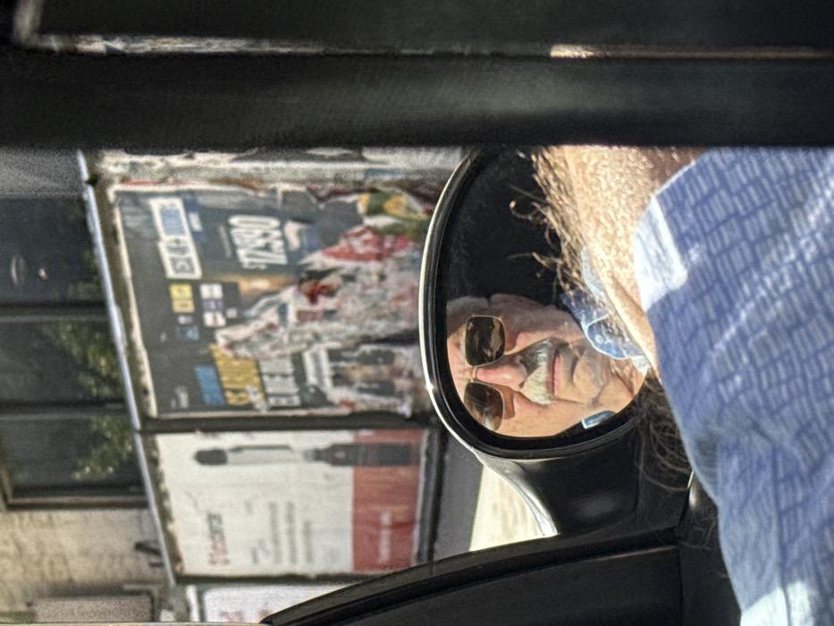 The image shows a car's side mirror reflecting an older man with sunglasses and a mustache. He's wearing a blue shirt. Outside the car, a wall with multiple posters is visible, including one with "$73590" printed on it. The scene is viewed from inside the car, with parts of the car frame in the foreground. The image suggests a moment captured while driving through an urban area.