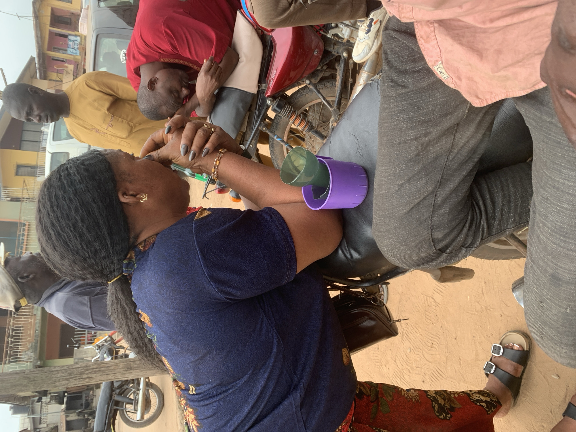 In Shagamu, Nigeria, a group of individuals are gathered around a motorcycle on a dirt road. A woman in a navy blue shirt and red patterned pants leans forward, her face obscured as she appears to be speaking with or comforting a man in a red shirt who is resting his head on his hands. To their left, a man in a yellow robe stands looking away. Behind them, another man in a grey shirt and a cap is visible. A motorcycle with a red body and black seat is central to the scene. A purple cup and a green funnel are placed on the motorcycle's seat. The environment suggests a street setting with buildings and other vehicles in the background.