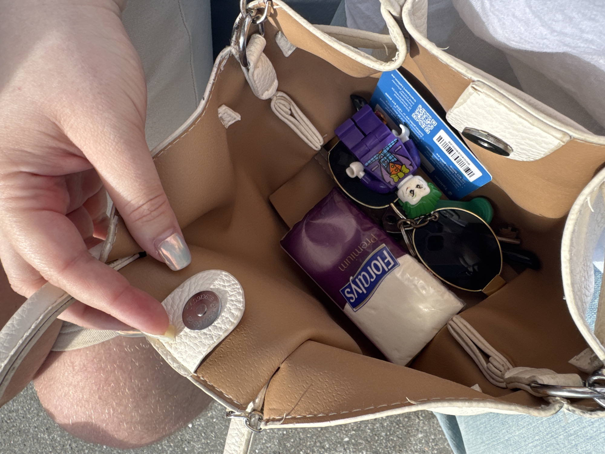 A person's hand, with a silver-toned nail polish, is reaching into an open, white, textured leather tote bag. The bag is lined with a tan-colored material and has silver hardware. Inside the bag are a pack of "Florals Premium" tissues, a set of keys with a green keychain that appears to be a cartoon character, and a purple Lego figure, likely a Joker. A blue and white card with a barcode and QR code is also visible, possibly a transit card or ticket. The background suggests an outdoor environment with sunlight and pavement, consistent with a street or public area in Tallinn, Estonia.